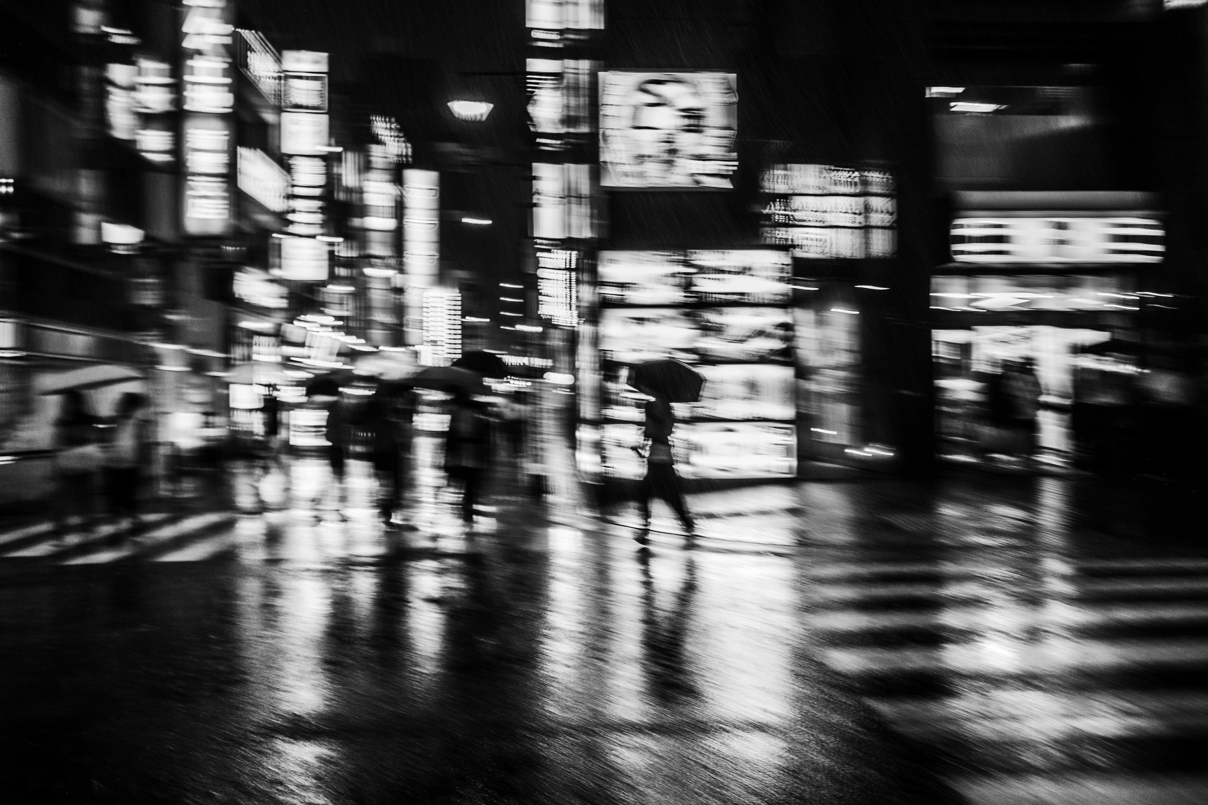 Night cityscape featuring people with umbrellas and bright signs reflecting on wet pavement