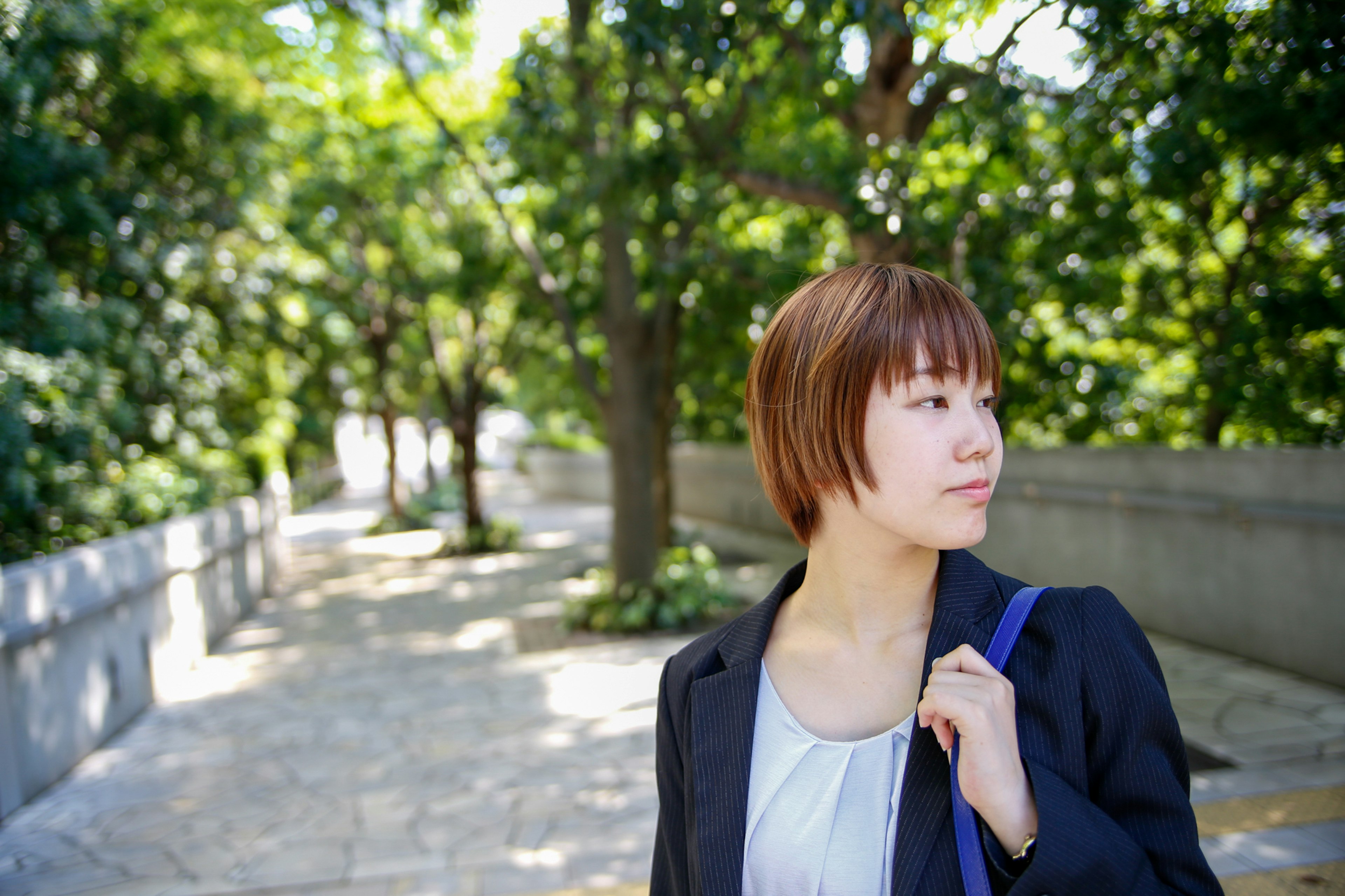 Young woman in profile at a park surrounded by green trees and stone path wearing a suit and holding a bag