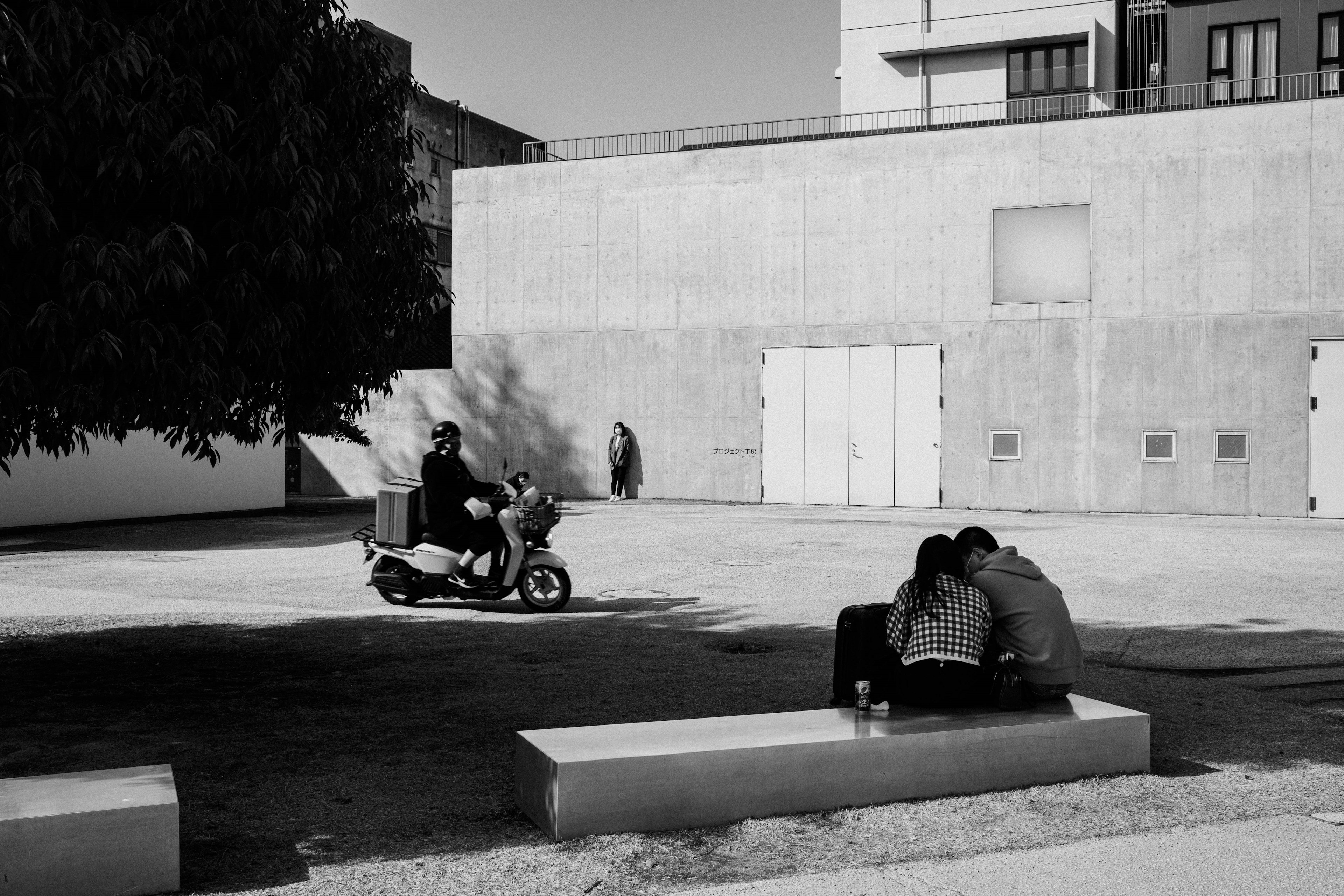 Image en noir et blanc d'un couple assis sur un banc dans un parc avec une personne conduisant un scooter