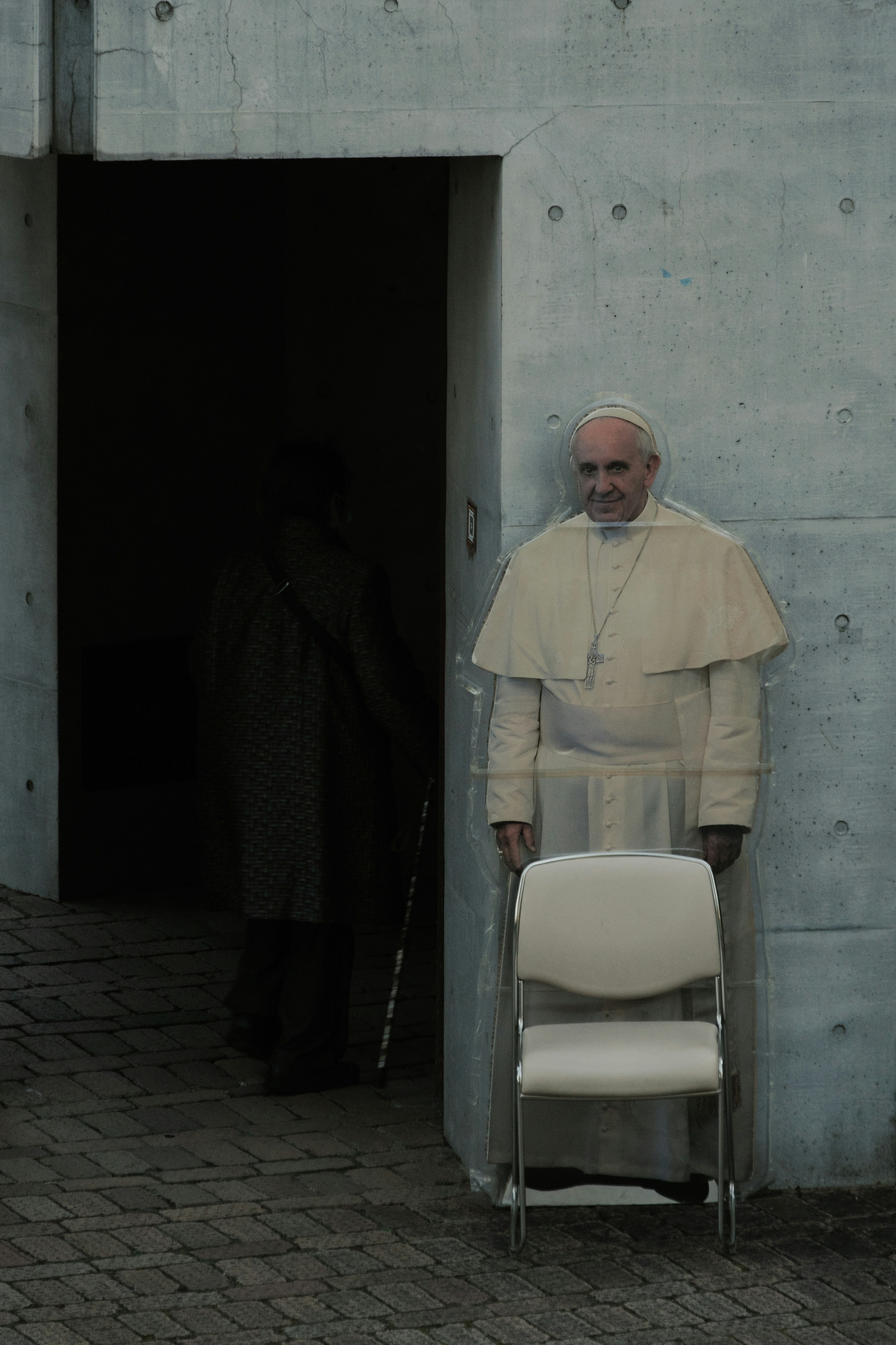 Pope standing in white robes next to a chair near a concrete wall with a dark entrance