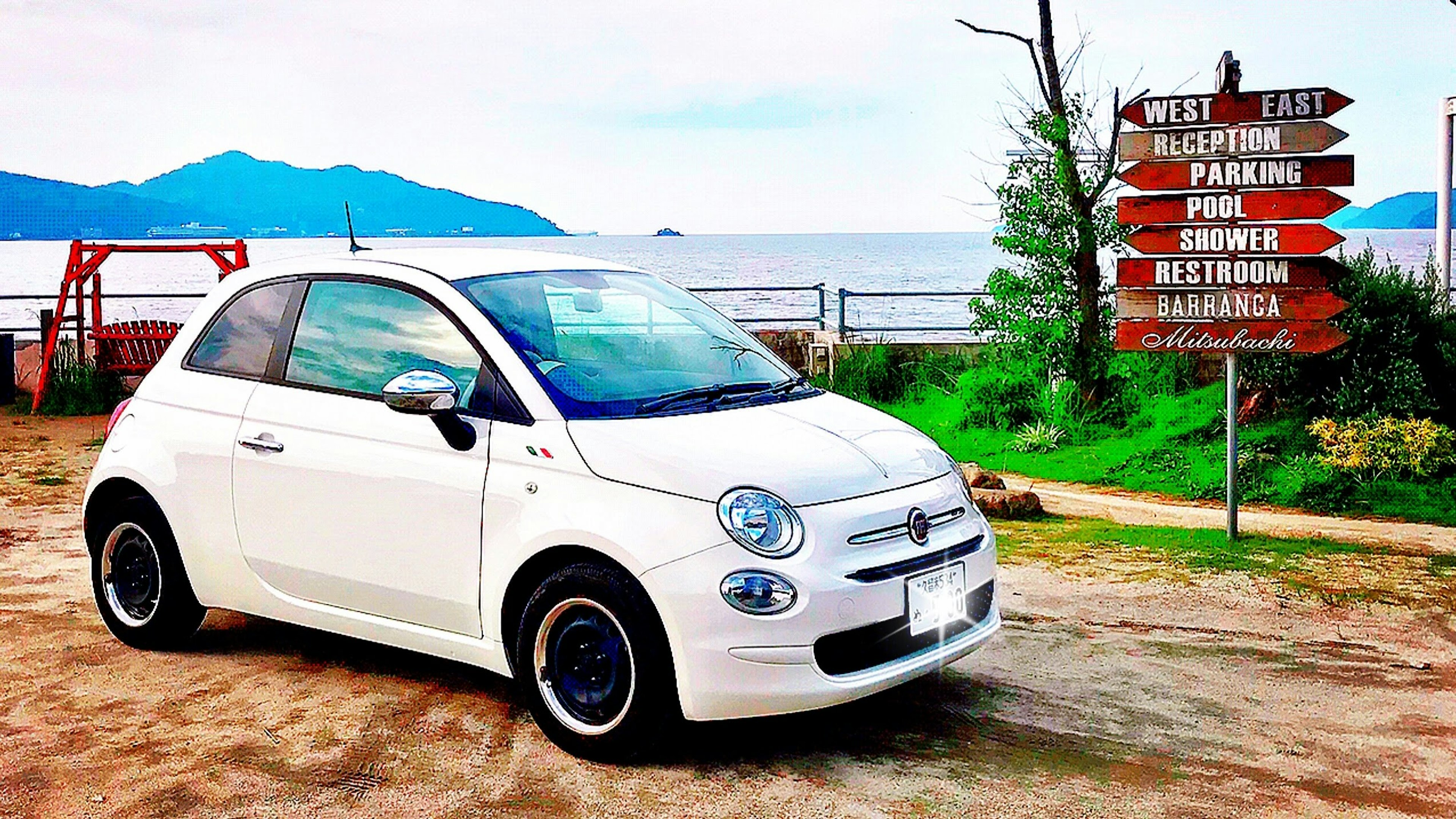 Sebuah Fiat 500 putih diparkir dekat pantai dengan tanda arah di latar belakang