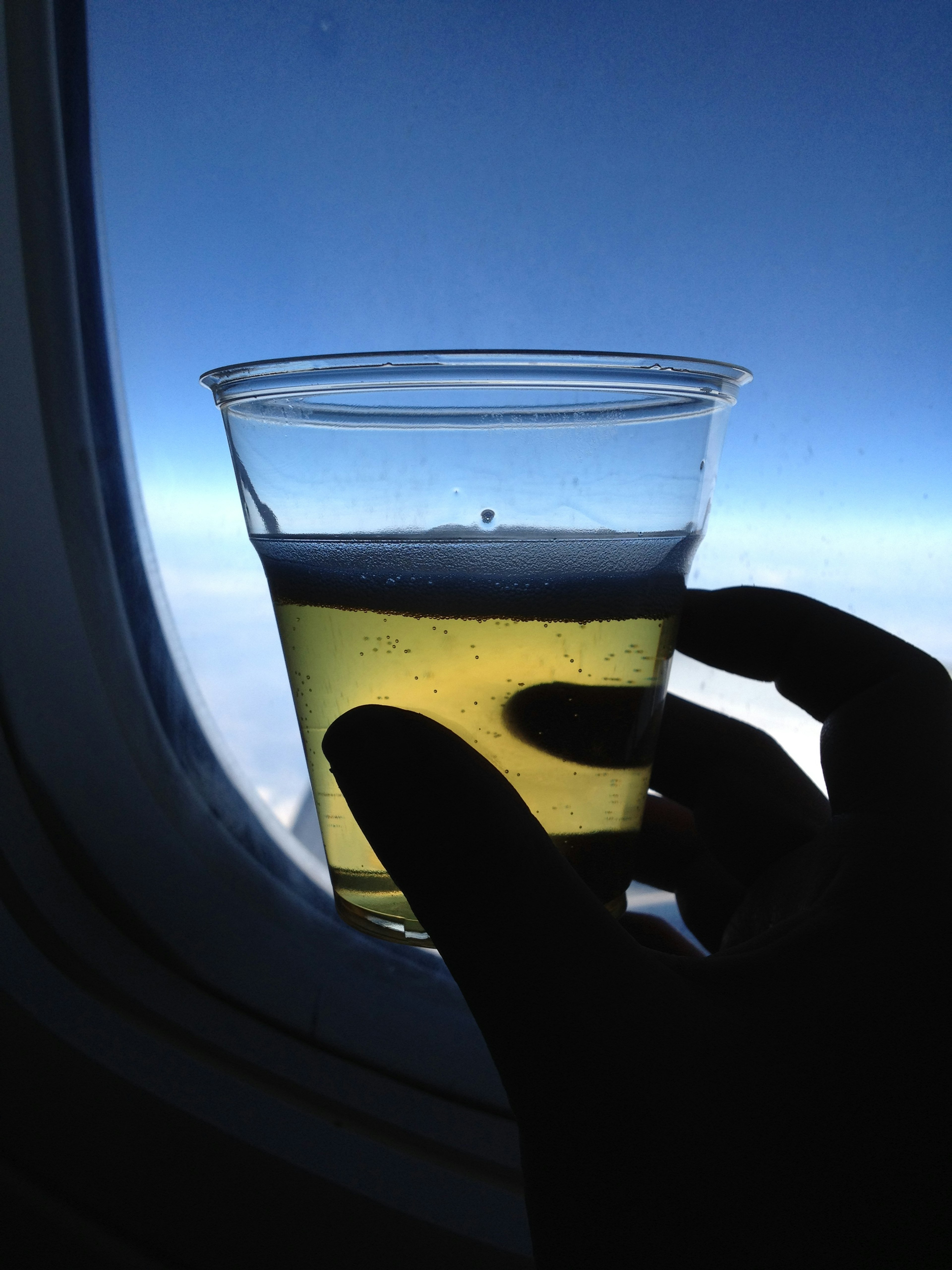 A hand holding a clear cup containing a yellow drink against a backdrop of blue sky outside an airplane window