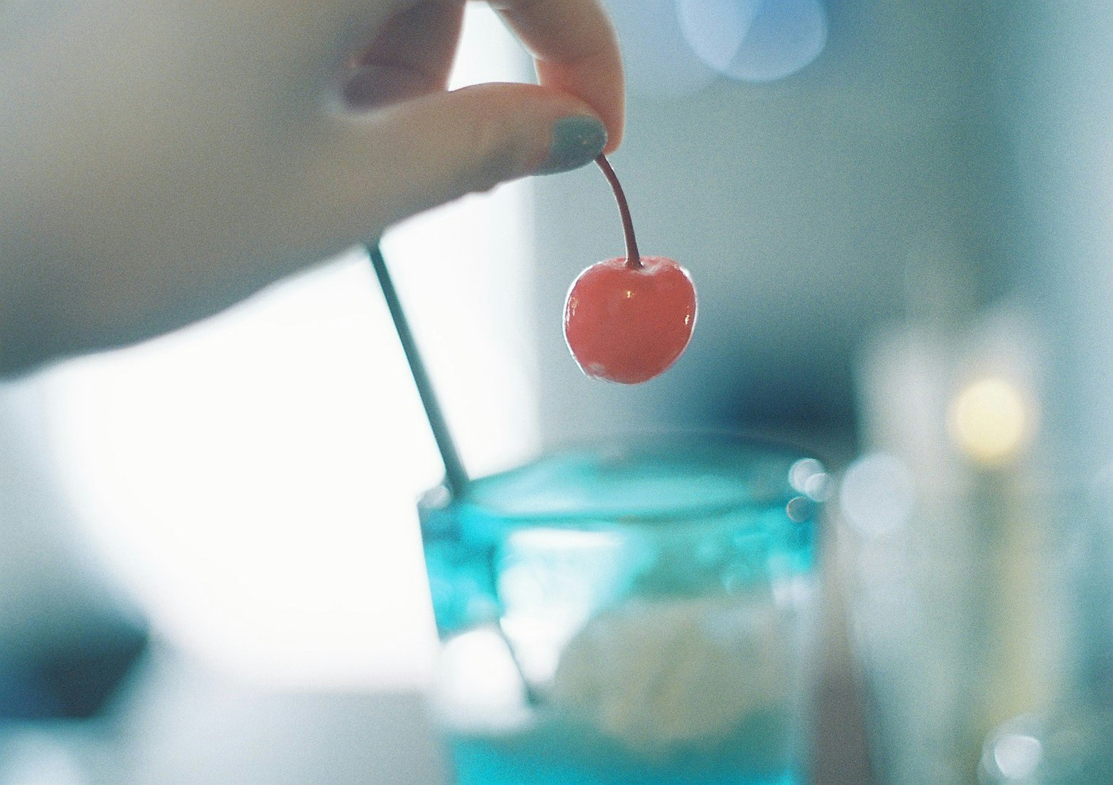 A hand holding a red cherry above a blue drink