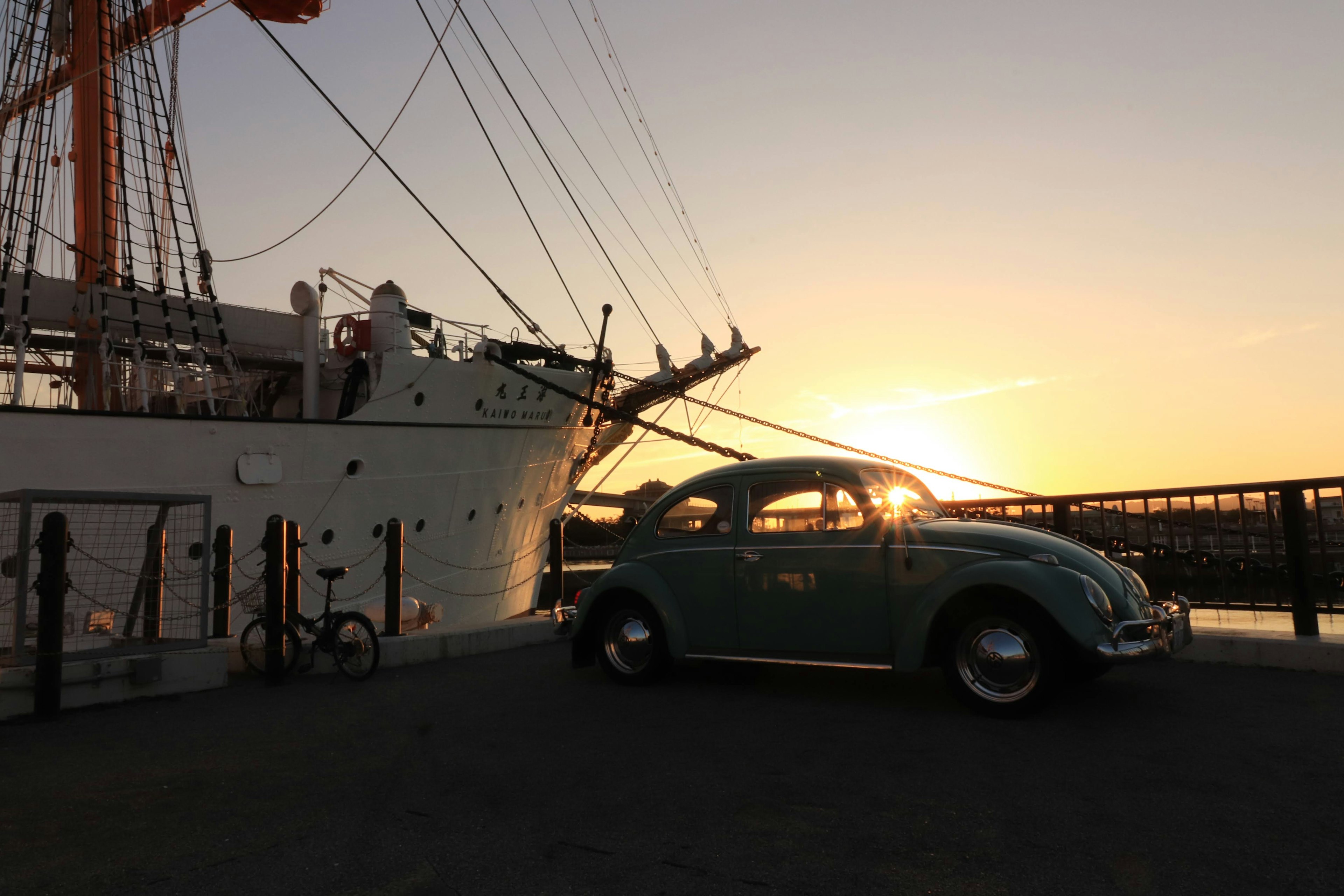 Vintage teal Volkswagen Beetle parked near a ship at sunset