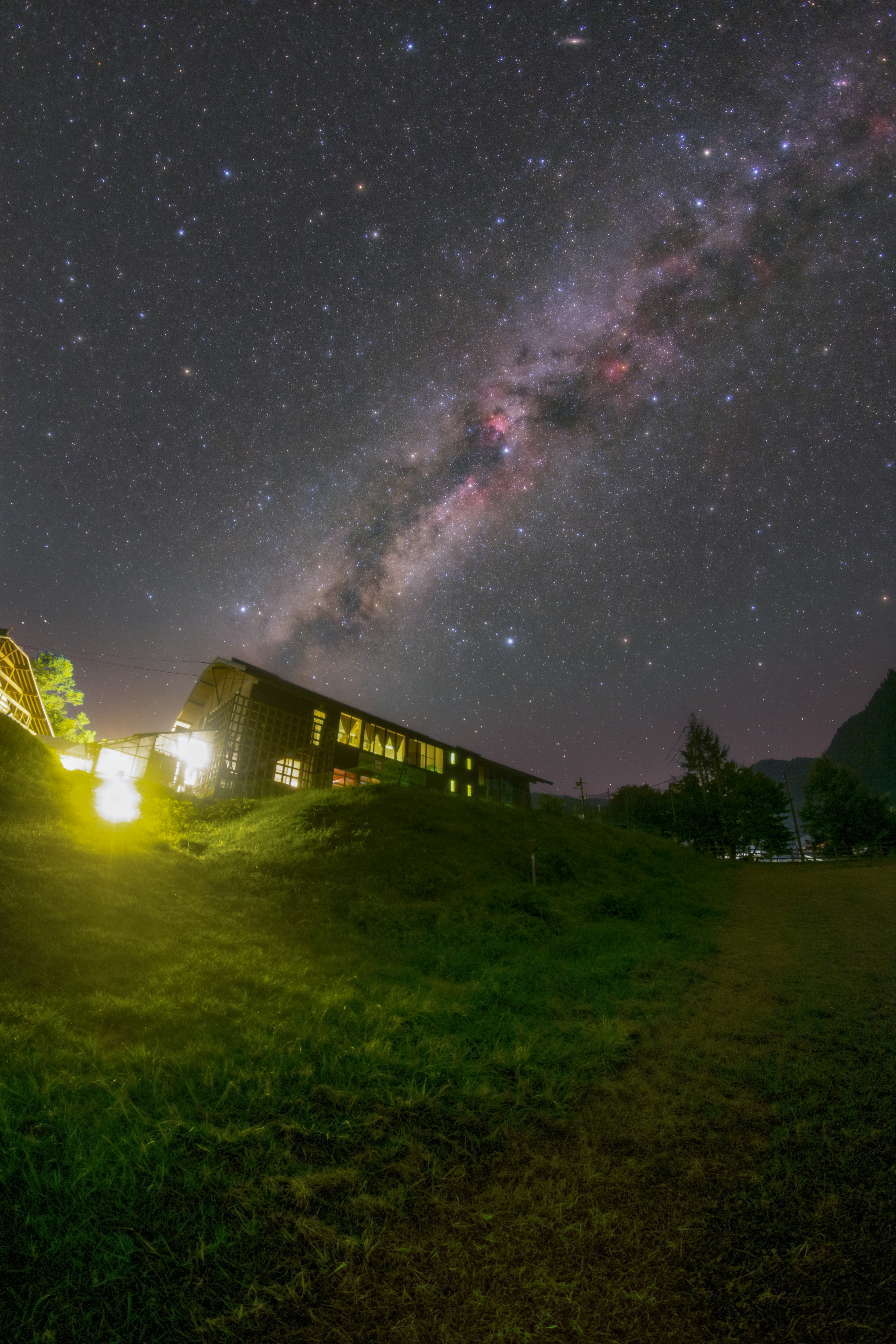 夜空に広がる天の川と緑の草地に立つ建物の光