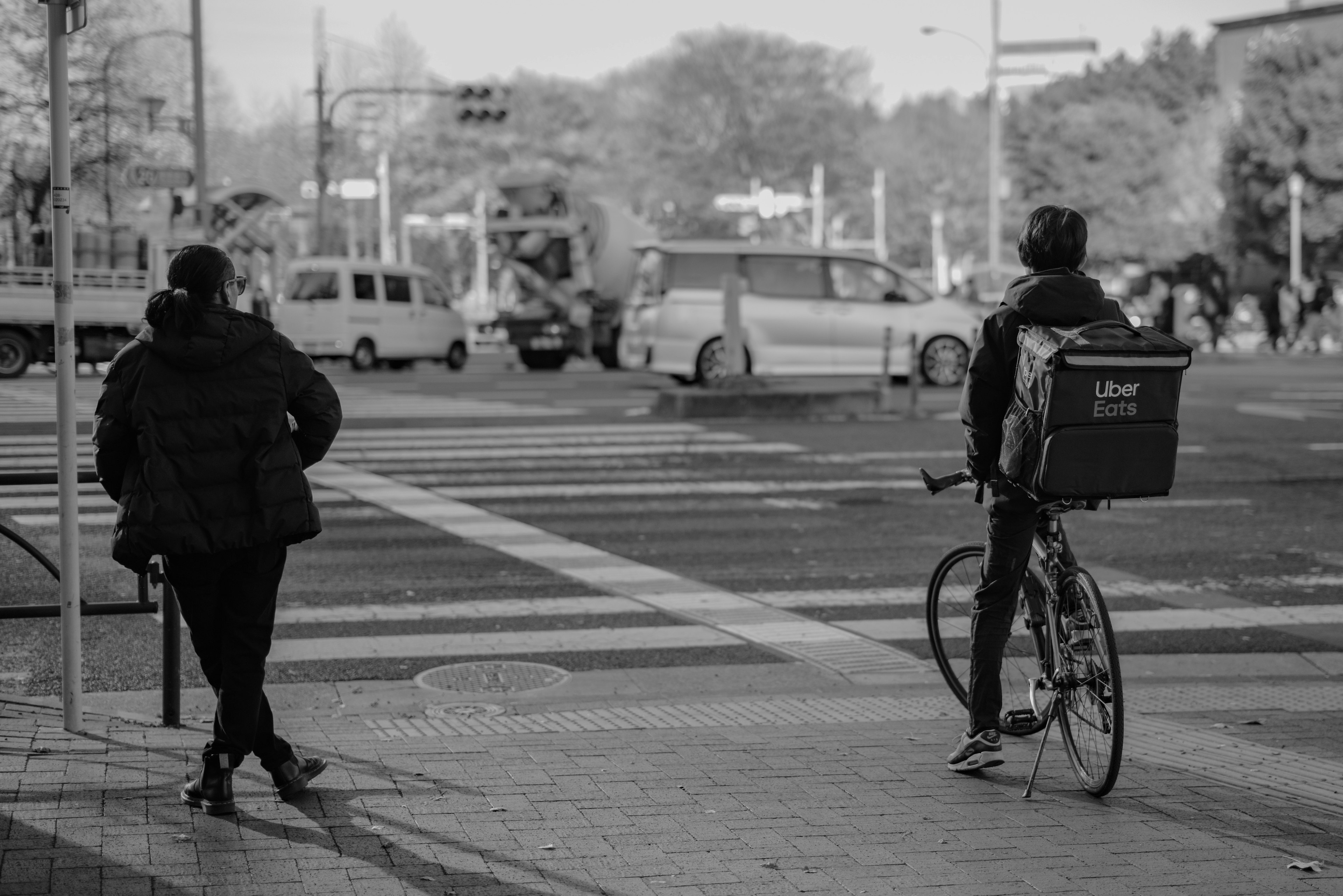 A cyclist with a delivery bag and a pedestrian waiting at a crosswalk in black and white