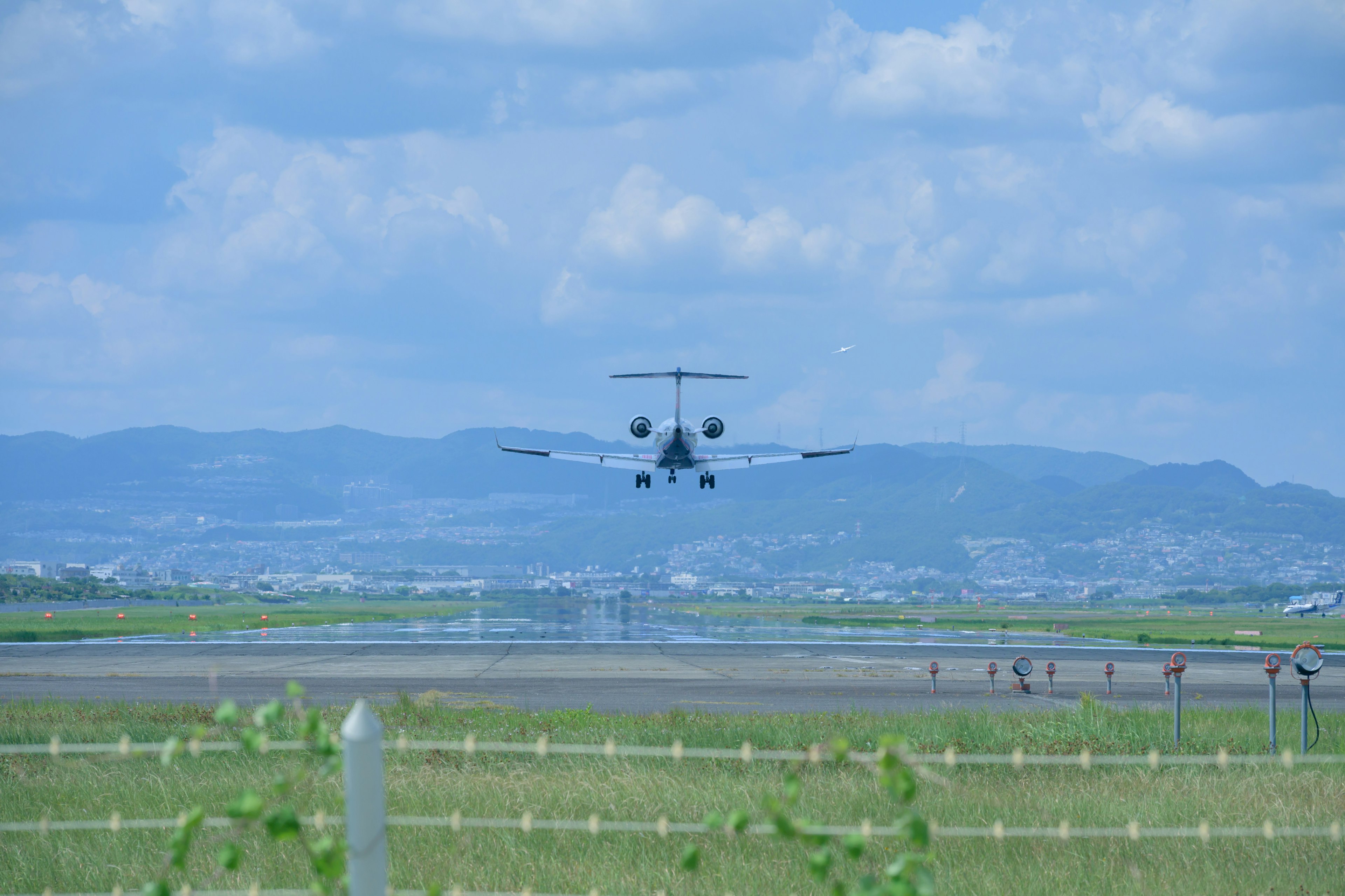 Un aereo in atterraggio su una pista con cielo blu e montagne sullo sfondo erba verde e recinzione visibili