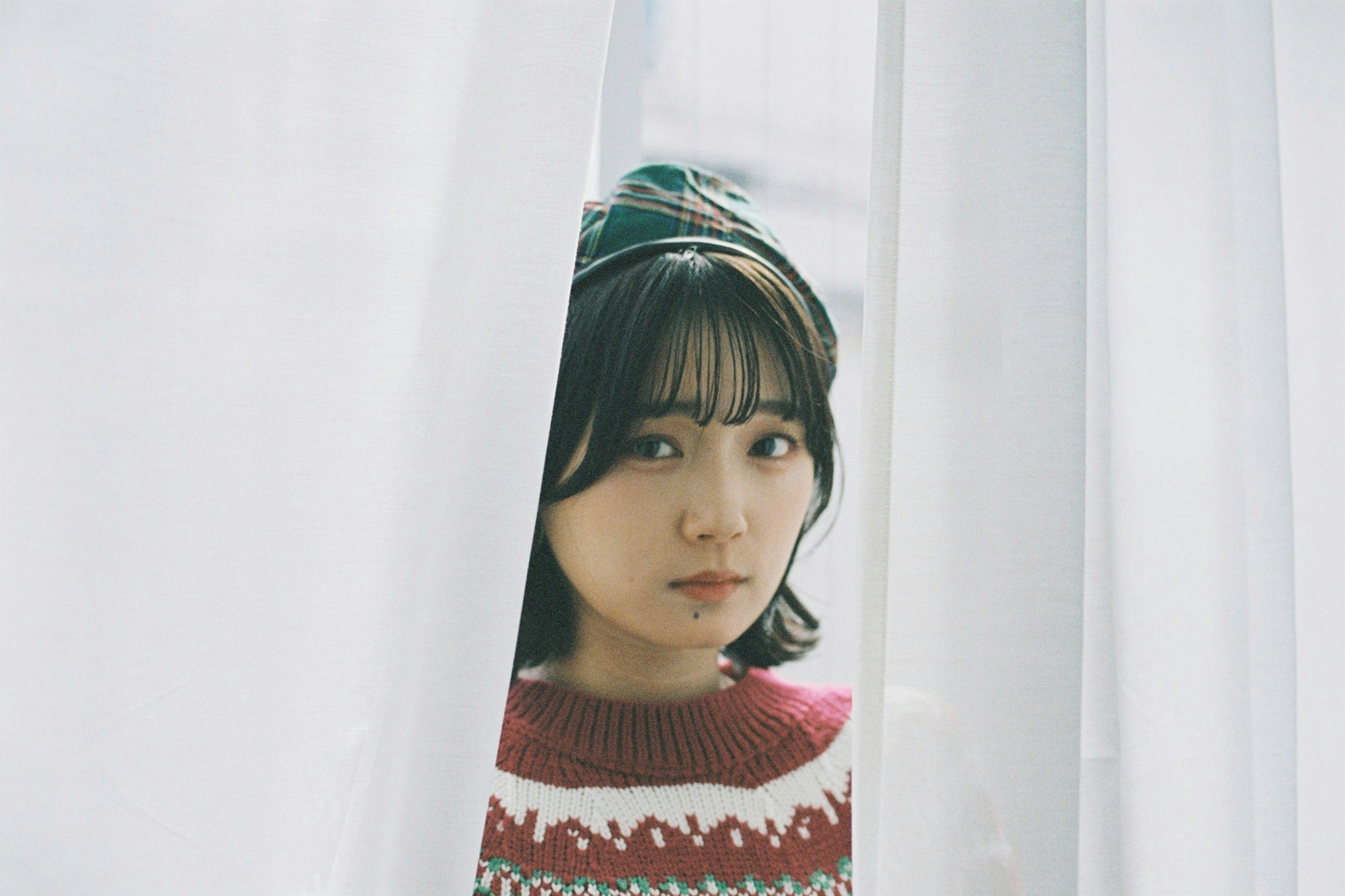 Portrait of a young woman peeking through white curtains wearing a red sweater