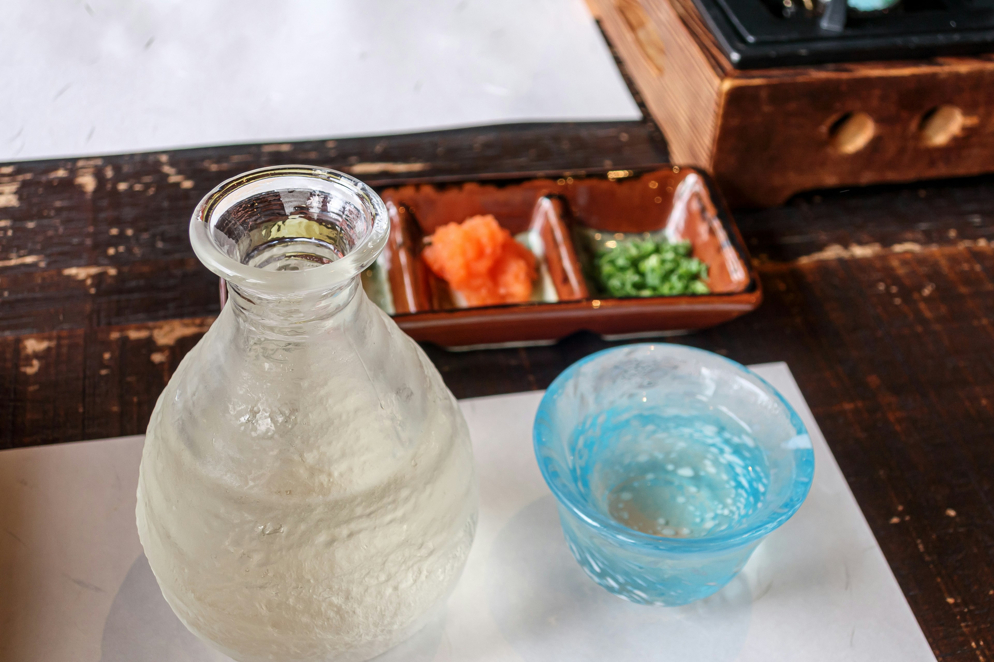 Botol sake bening dengan piring biru berisi es dan bumbu berwarna di samping