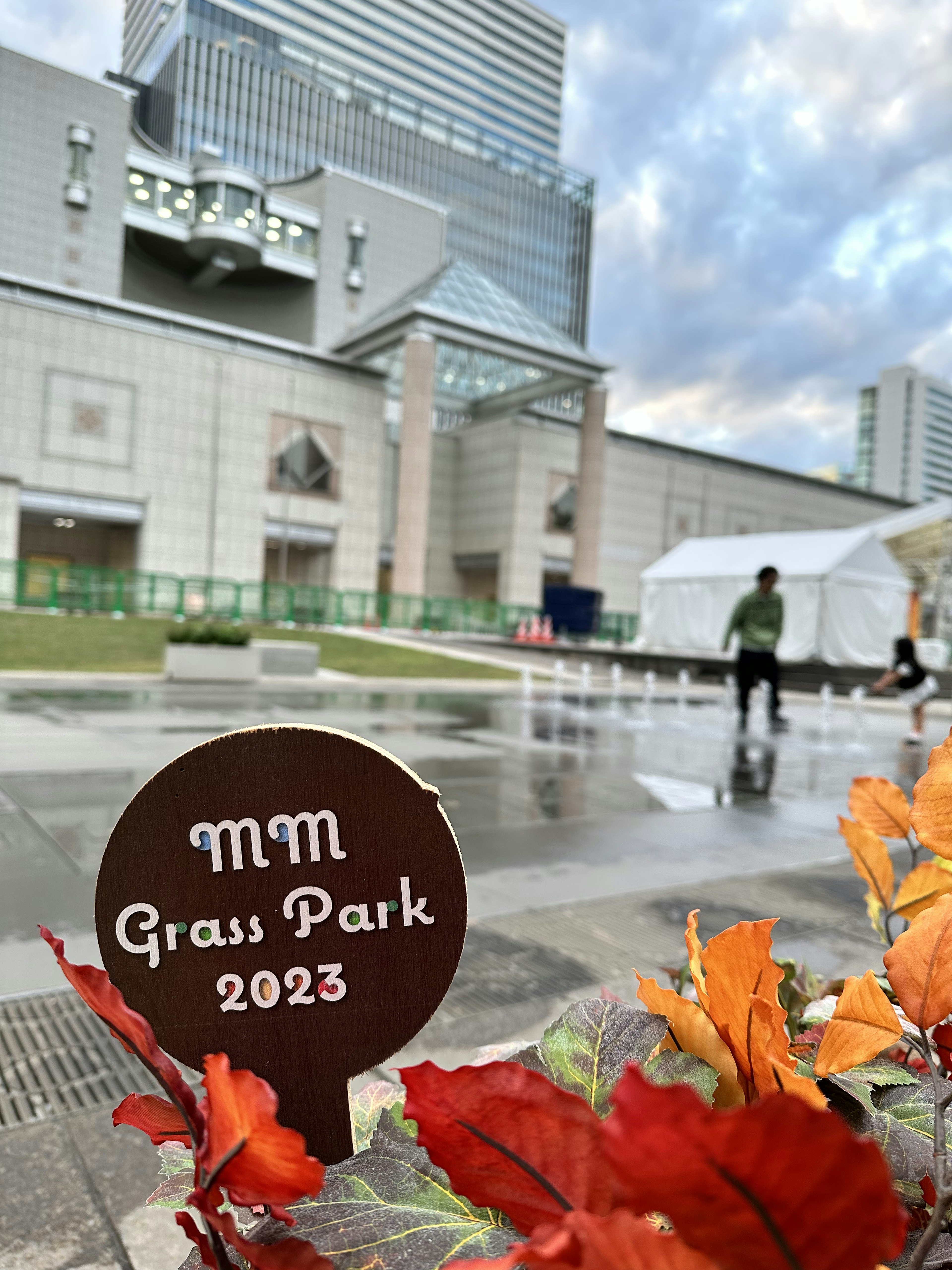 ทัศนียภาพของป้าย MM Grass Park 2023 โดยมีอาคารสมัยใหม่และใบไม้ฤดูใบไม้ร่วงอยู่เบื้องหลัง