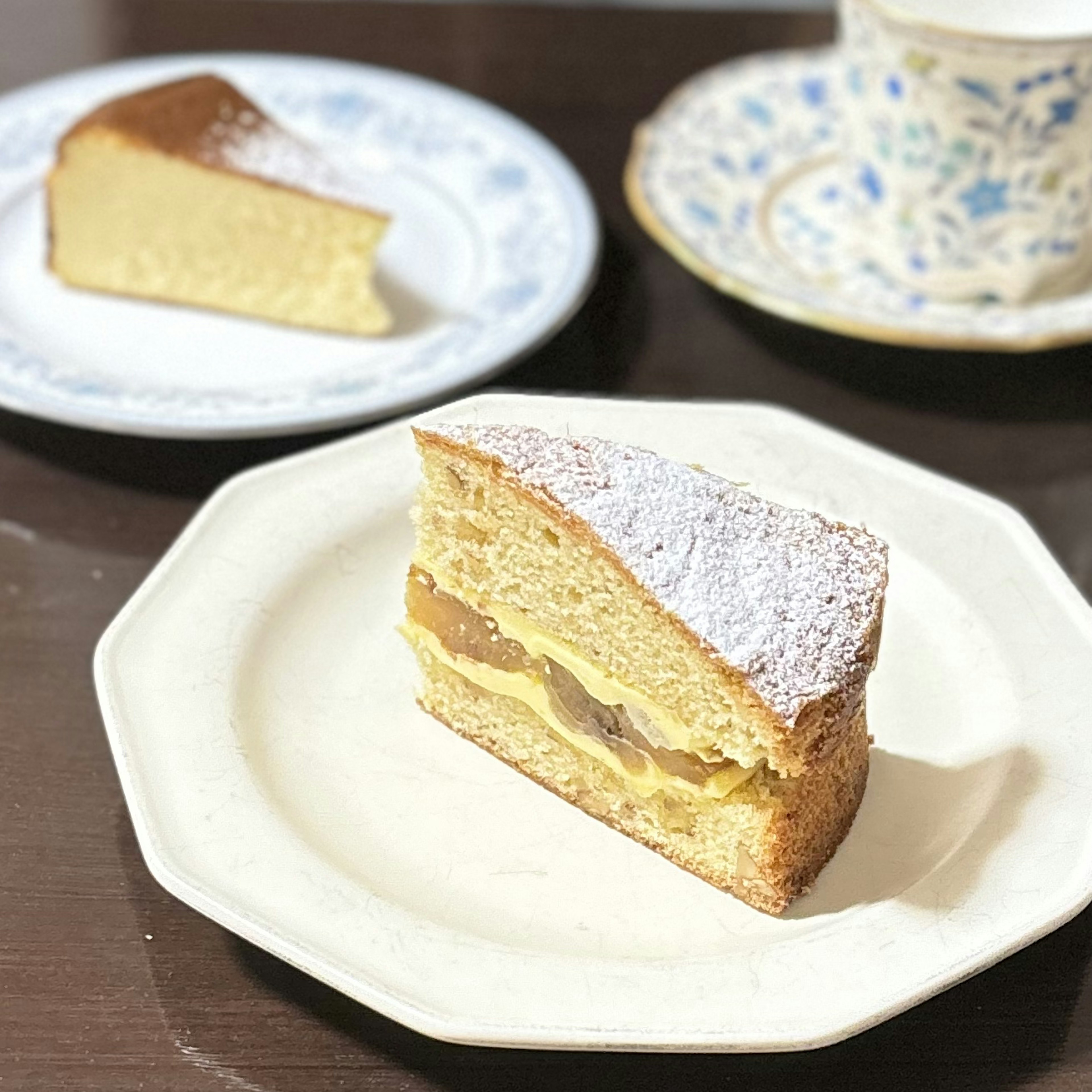 Ein Stück Biskuitkuchen auf einem dekorativen Teller mit einer Teetasse im Hintergrund