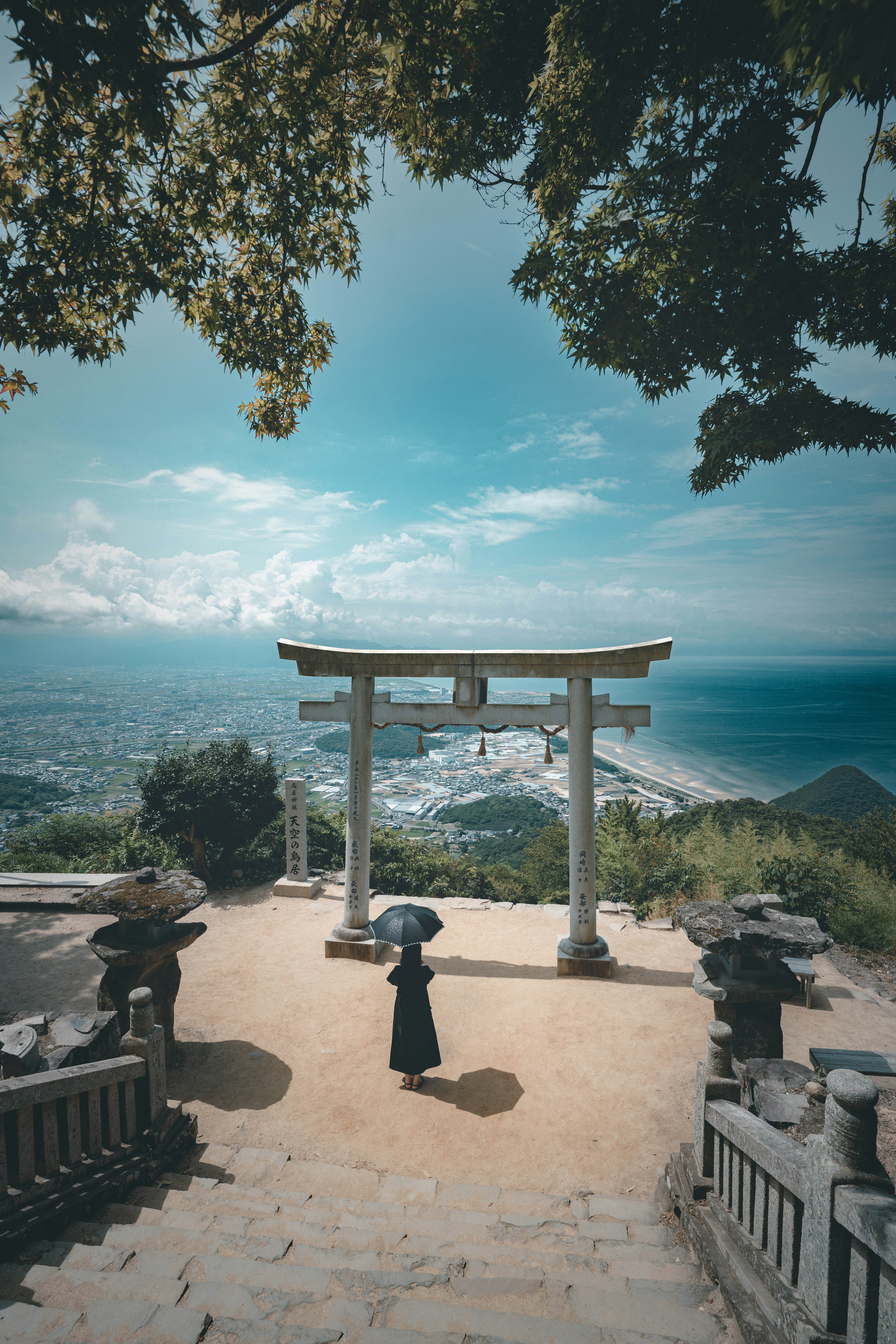 Eine Person in schwarzer Kleidung steht unter einem Torii mit Blick auf den Ozean und den blauen Himmel