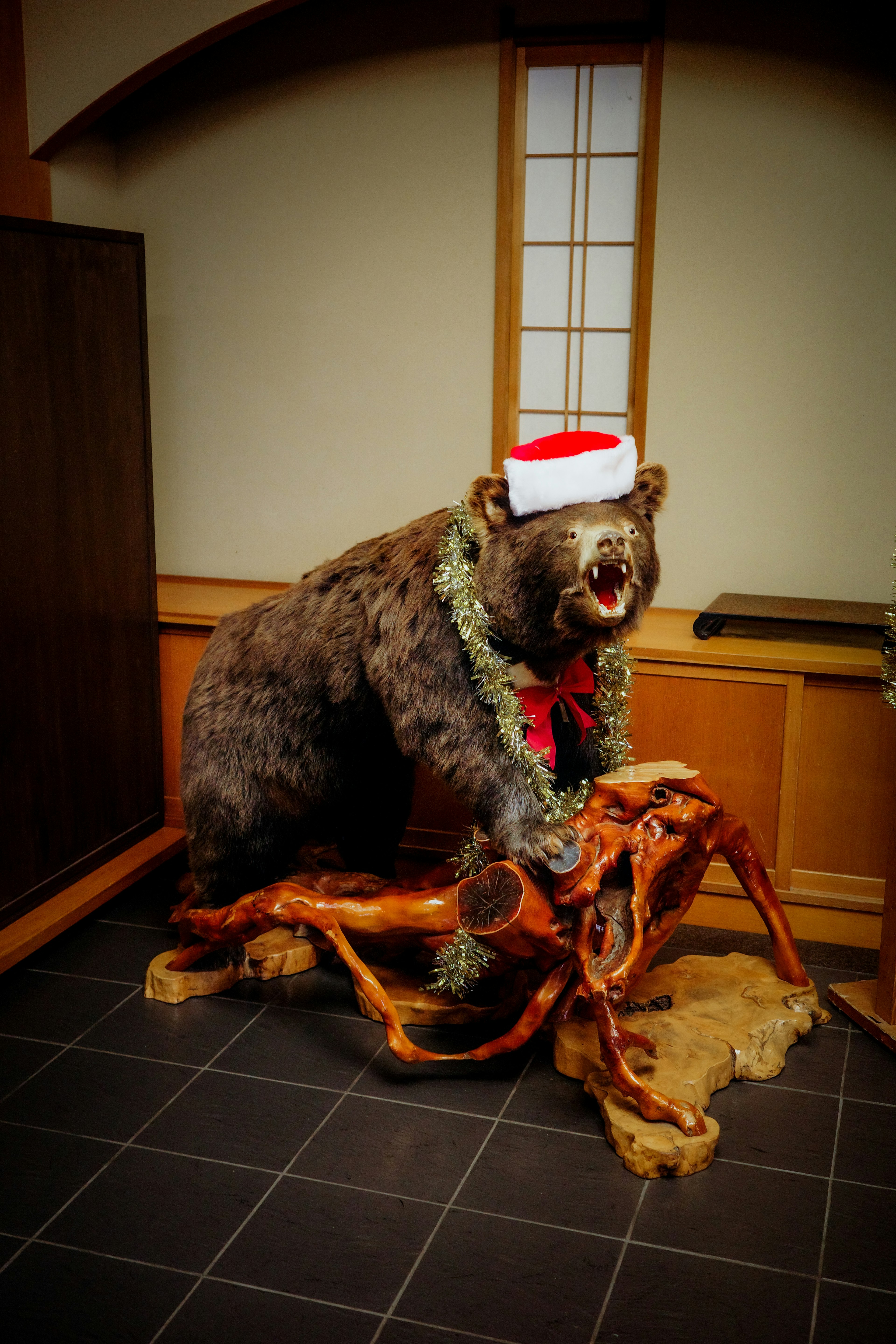 Ein ausgestopfter Bär, der einen Weihnachtsmannhut mit rotem Band und Girlande trägt, steht auf einer Holzbasis