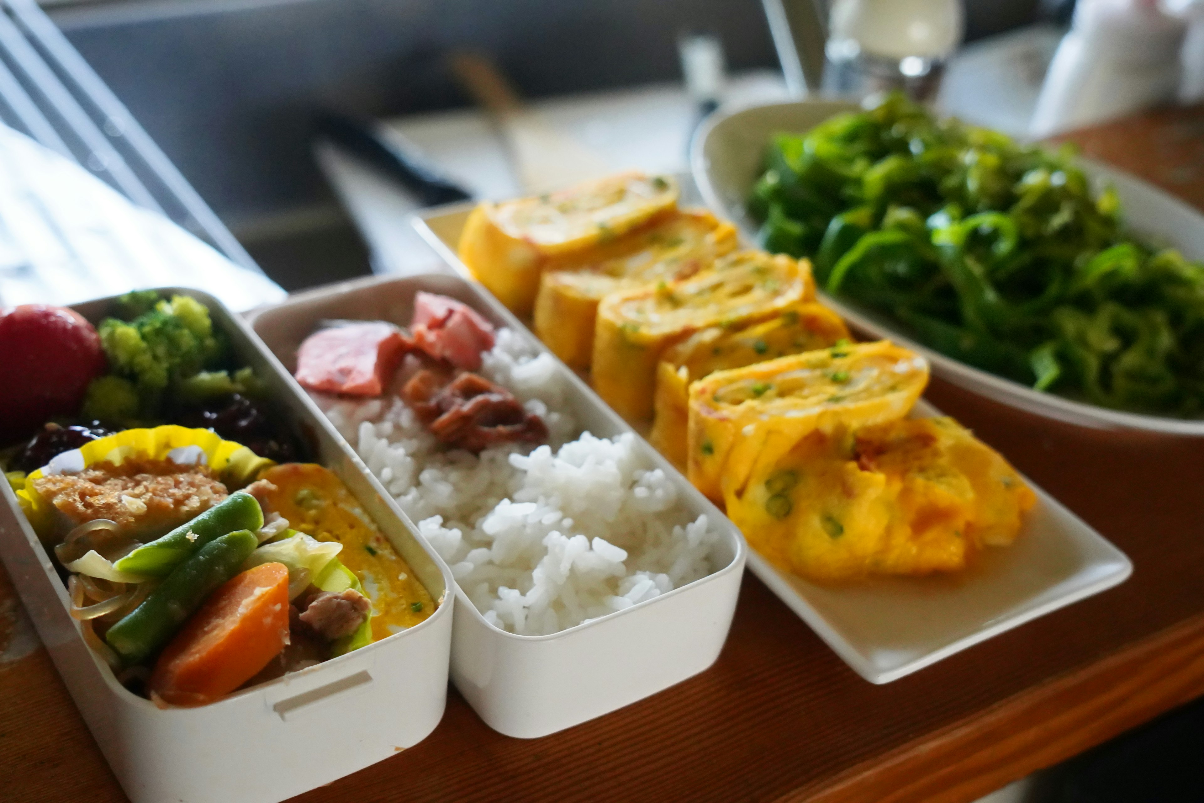 Kotak bento berwarna-warni dengan nasi sayuran dan omelet gulung di samping salad segar