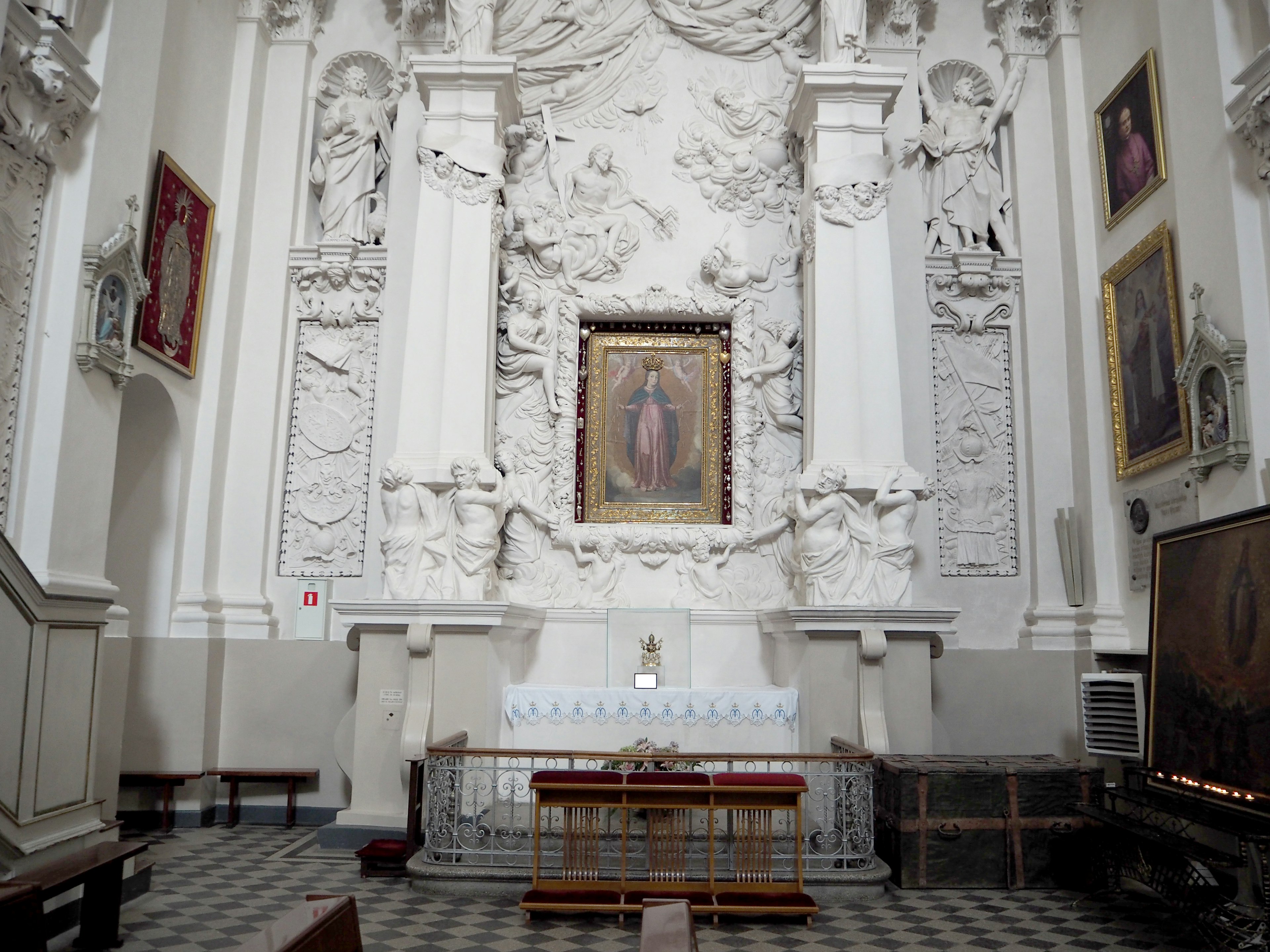 Interior of a church with white walls and intricate sculptures featuring a painting of the Virgin Mary on the altar