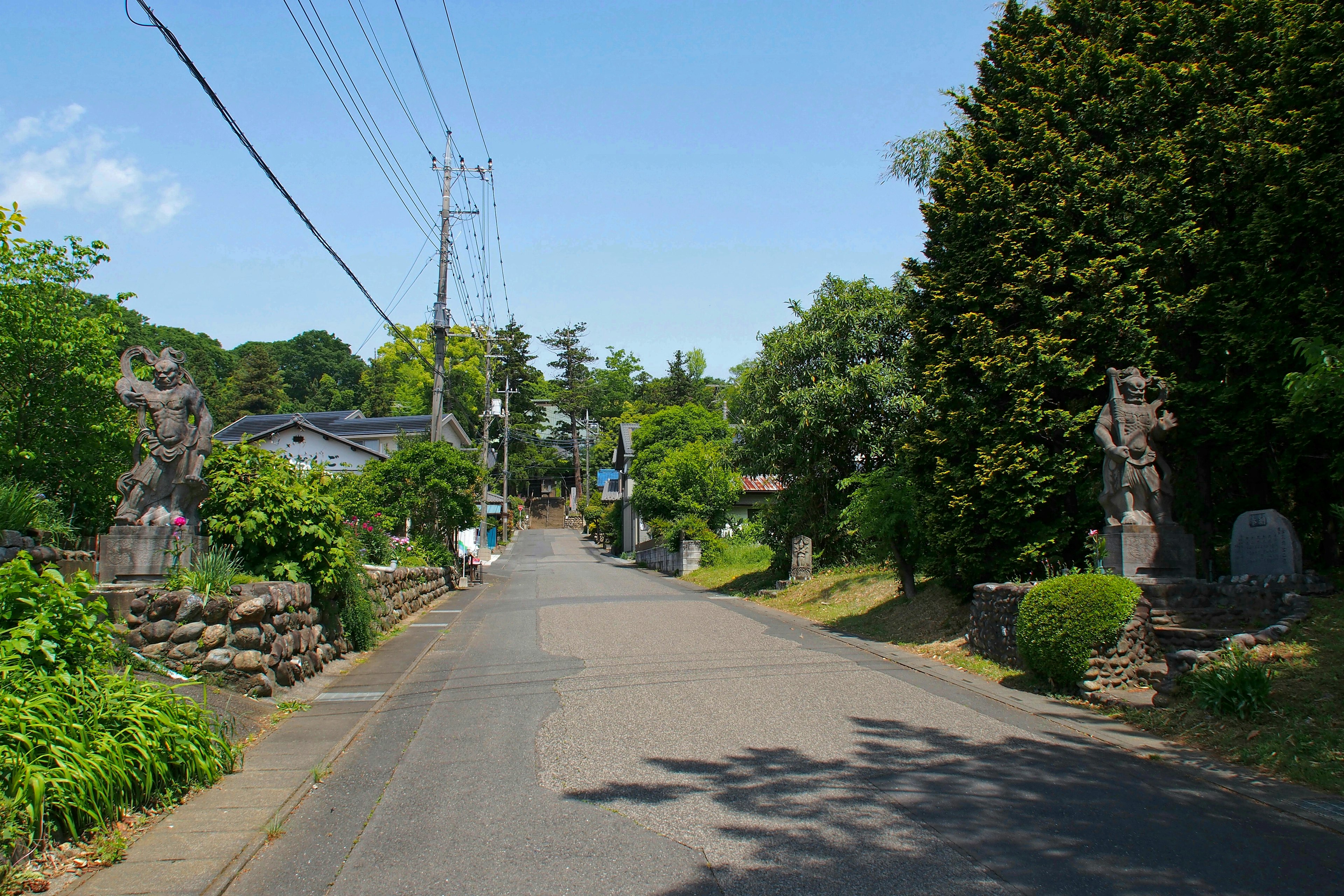 Quiet street lined with greenery and stone statues under a clear blue sky