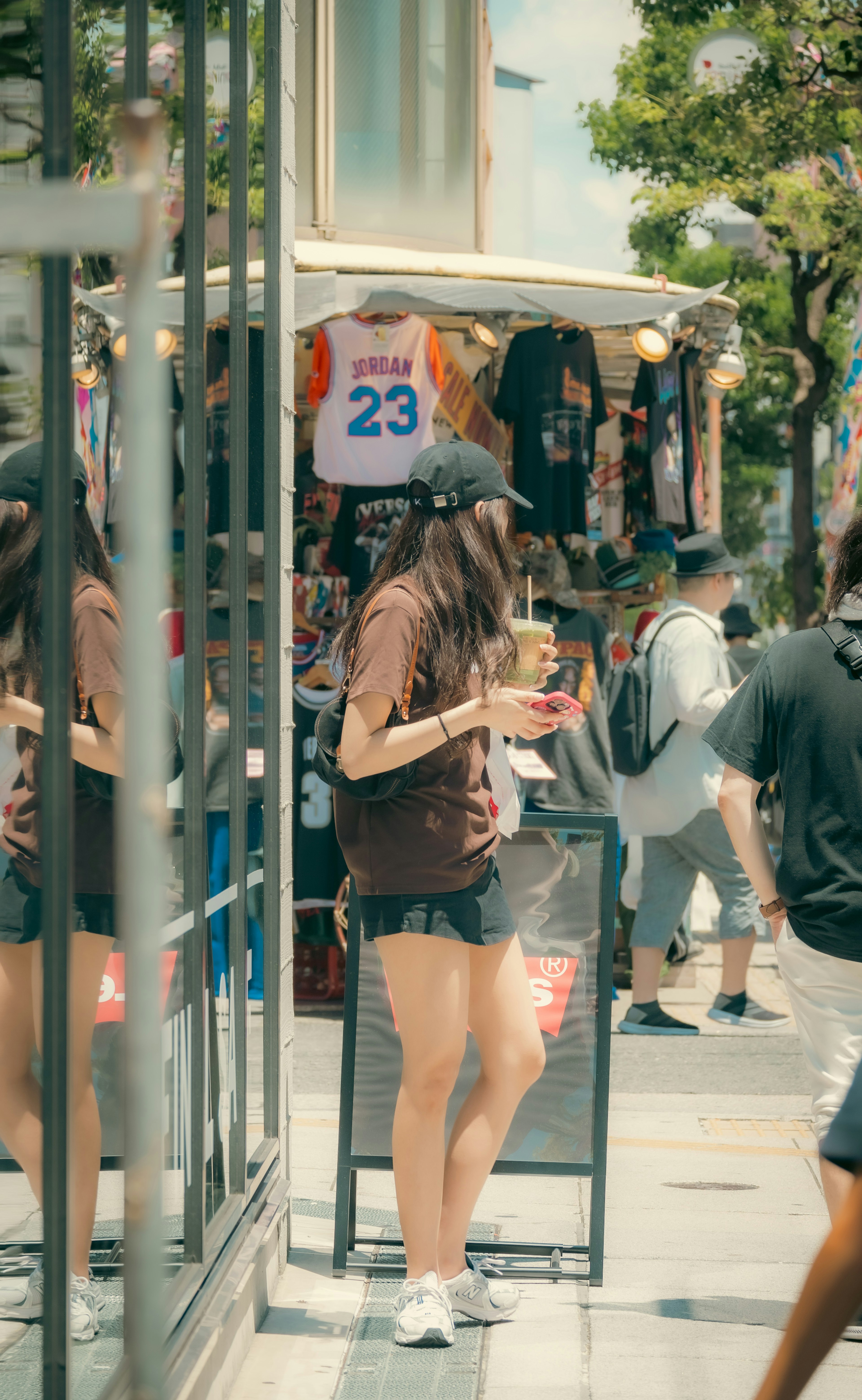 Young woman holding a drink standing in front of a shop with T-shirts displayed in the background