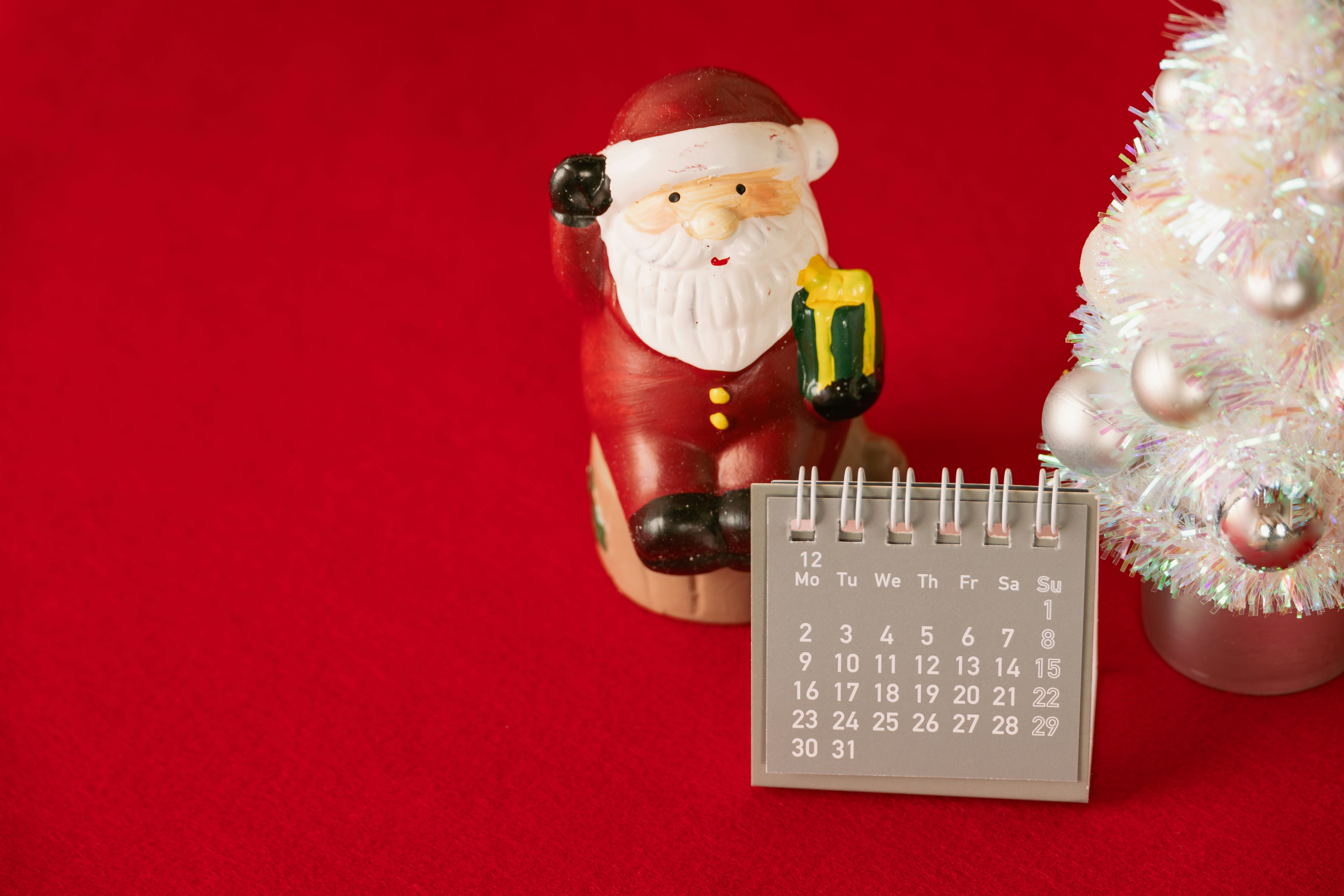 Figurine Santa Claus memegang objek kuning di sebelah pohon Natal putih kecil dan kalender di latar belakang merah