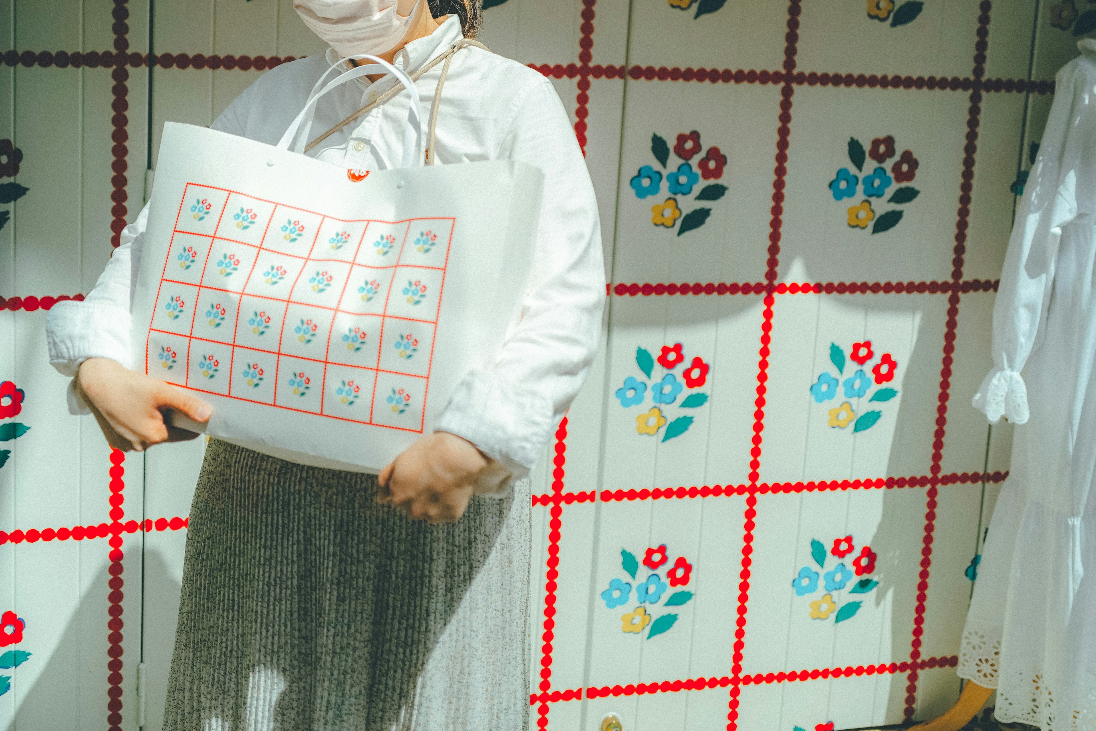 A woman in a white outfit holding a tote bag in front of a floral patterned background