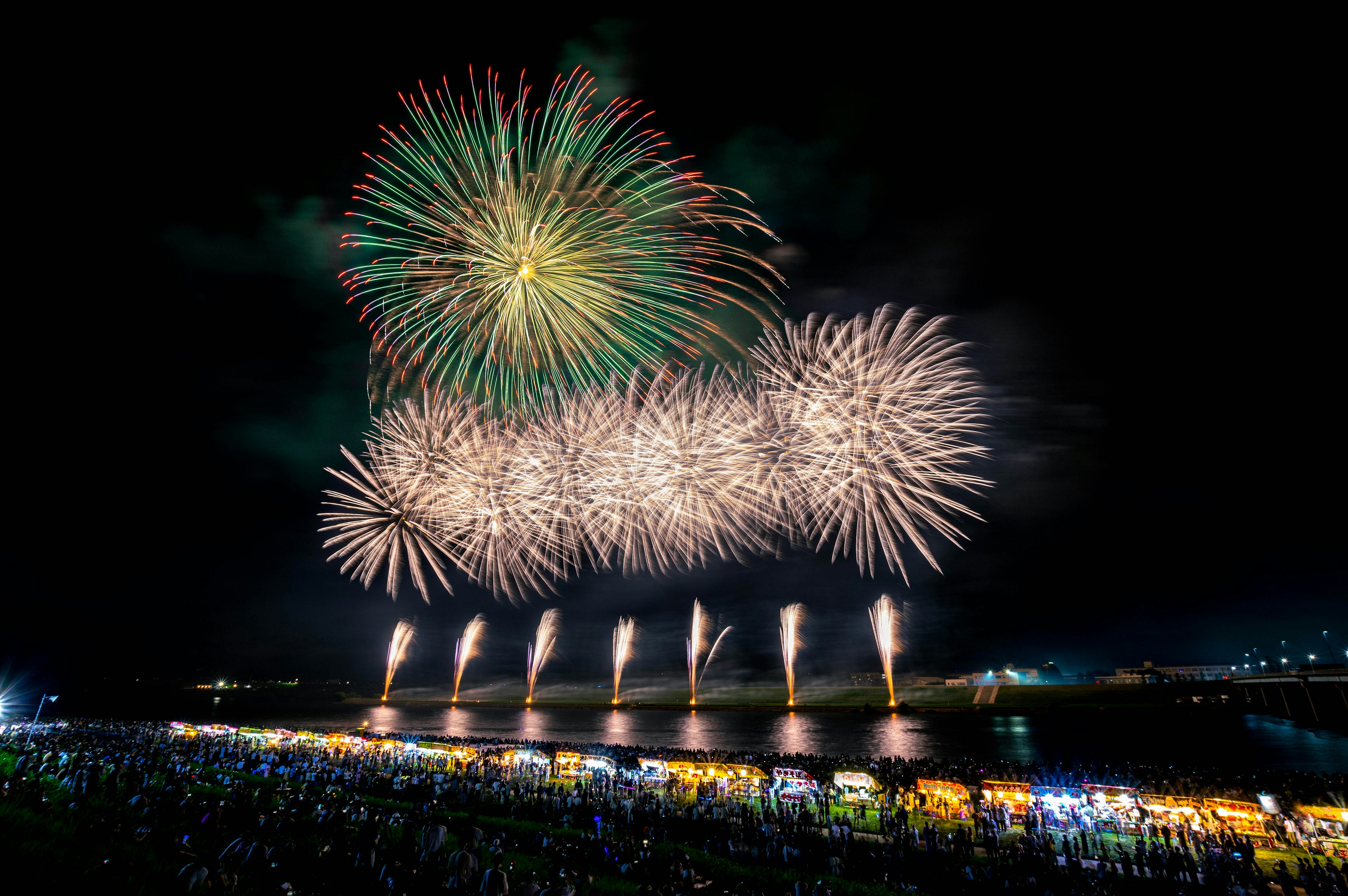 Colorful fireworks illuminate the night sky above a river with a large crowd watching