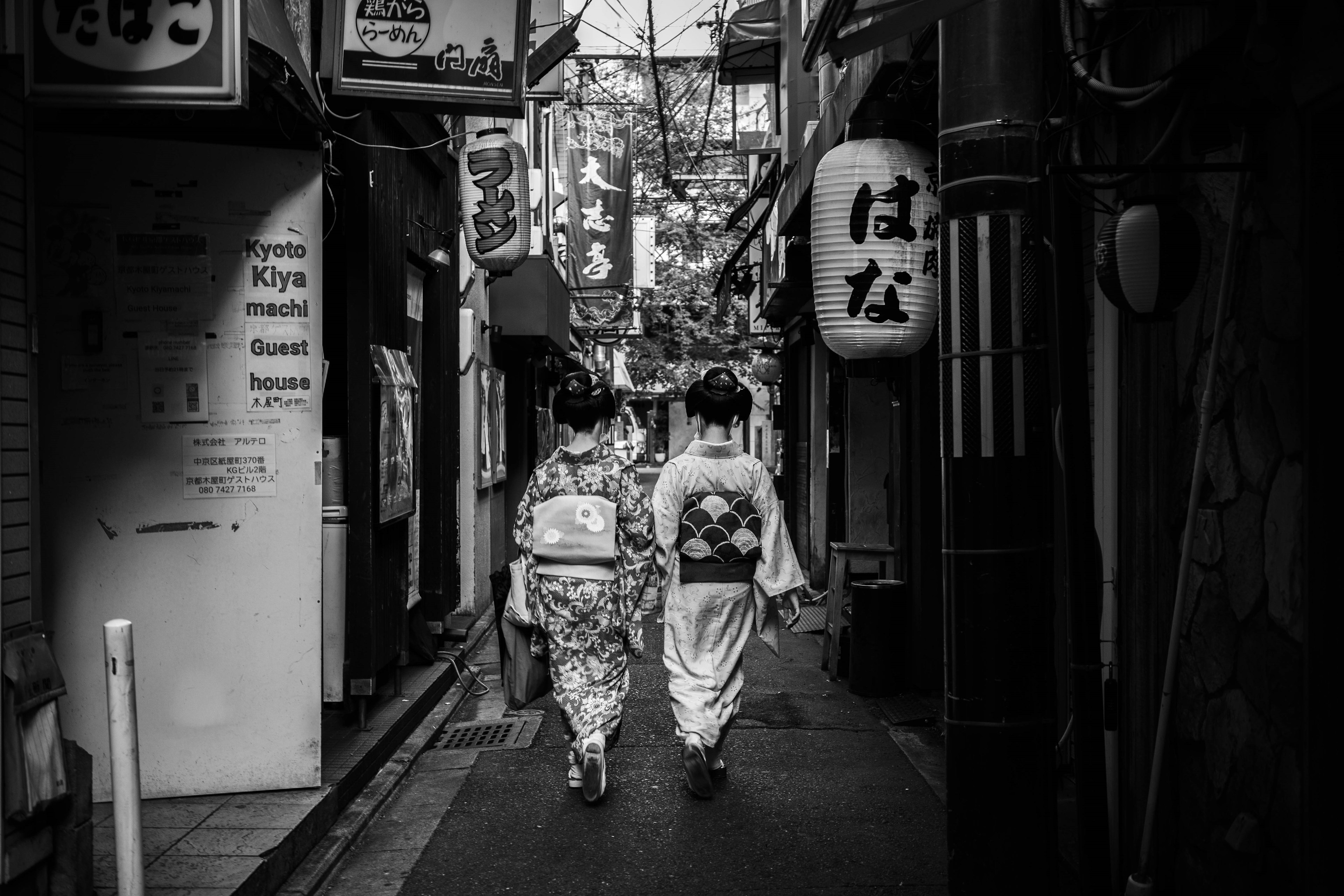 Two women in traditional kimonos walking down a narrow alley in black and white