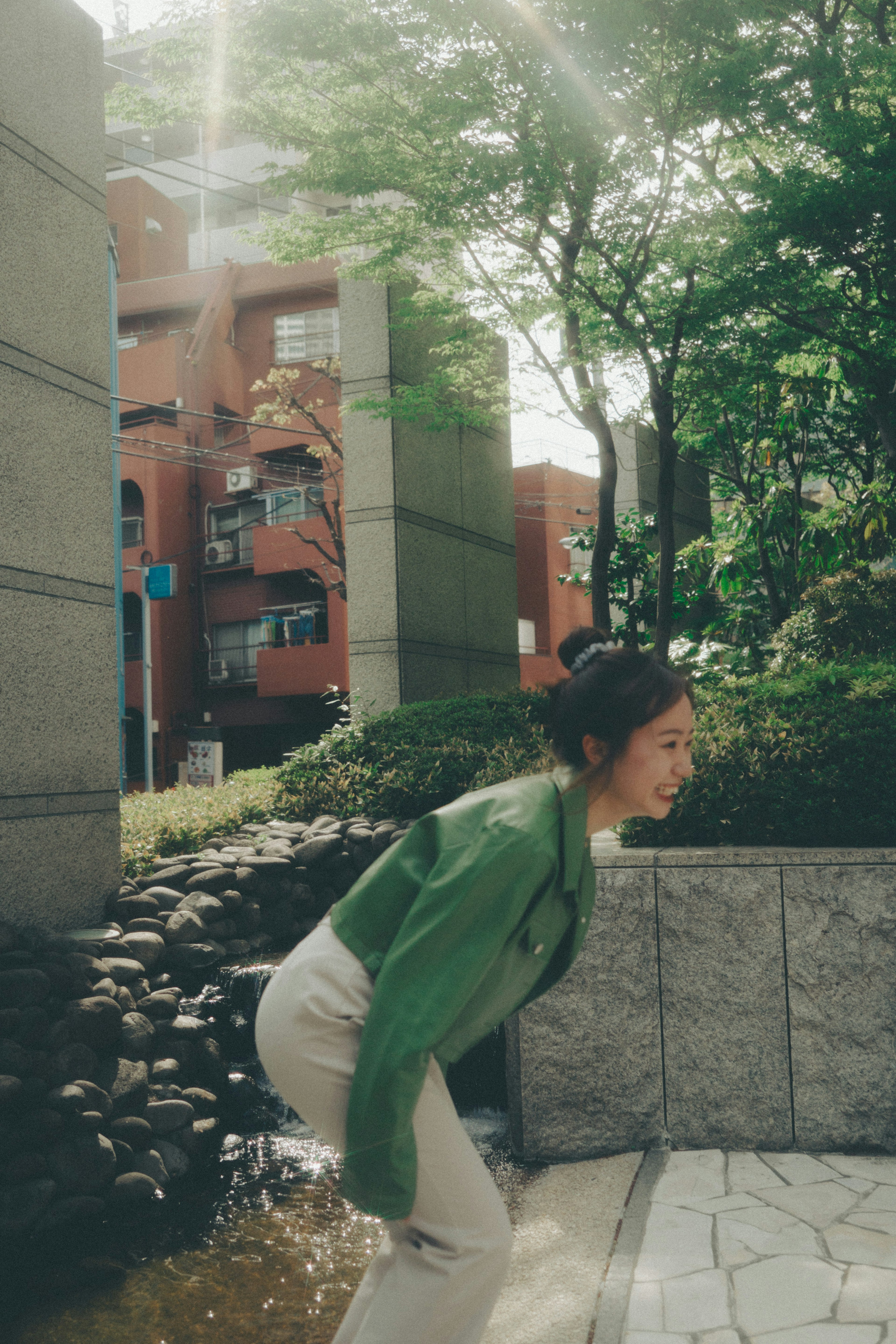 A joyful woman laughing in a park wearing a green jacket surrounded by trees and buildings