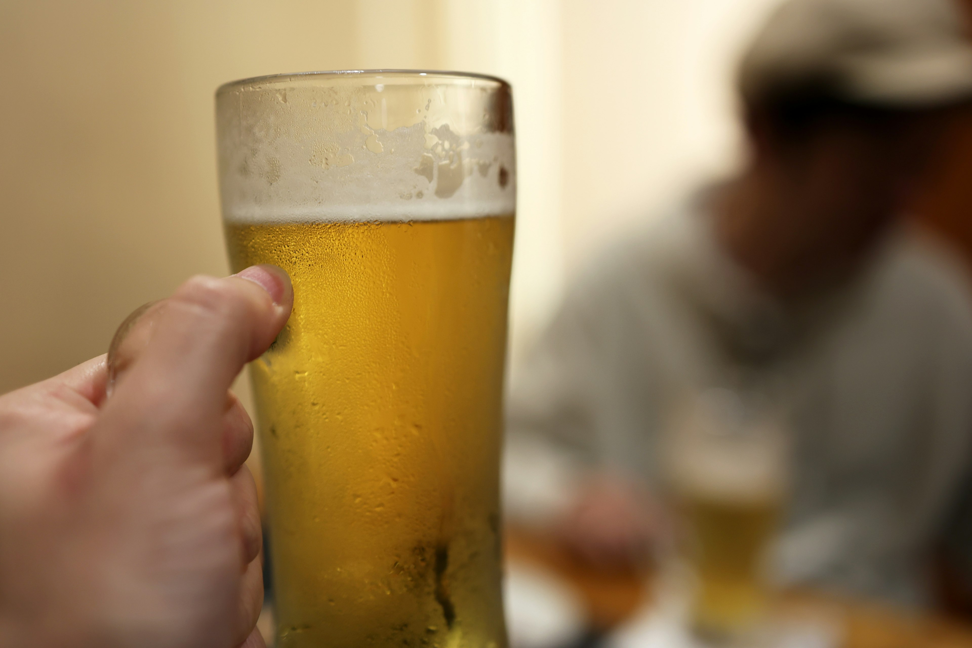 Hand hält ein Glas Bier mit einer verschwommenen Figur im Hintergrund