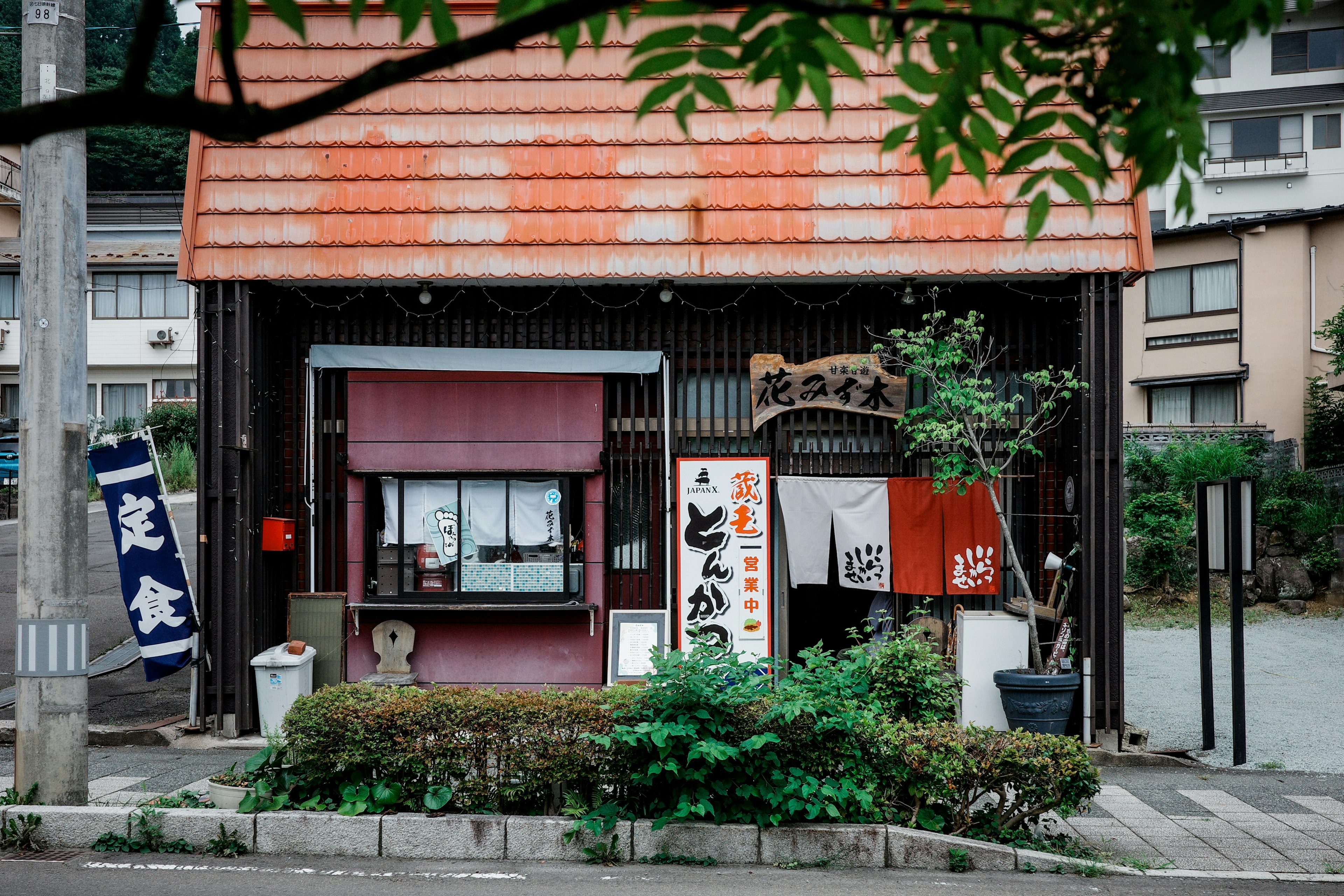 Small traditional Japanese eatery exterior with a red roof and banners surrounded by greenery