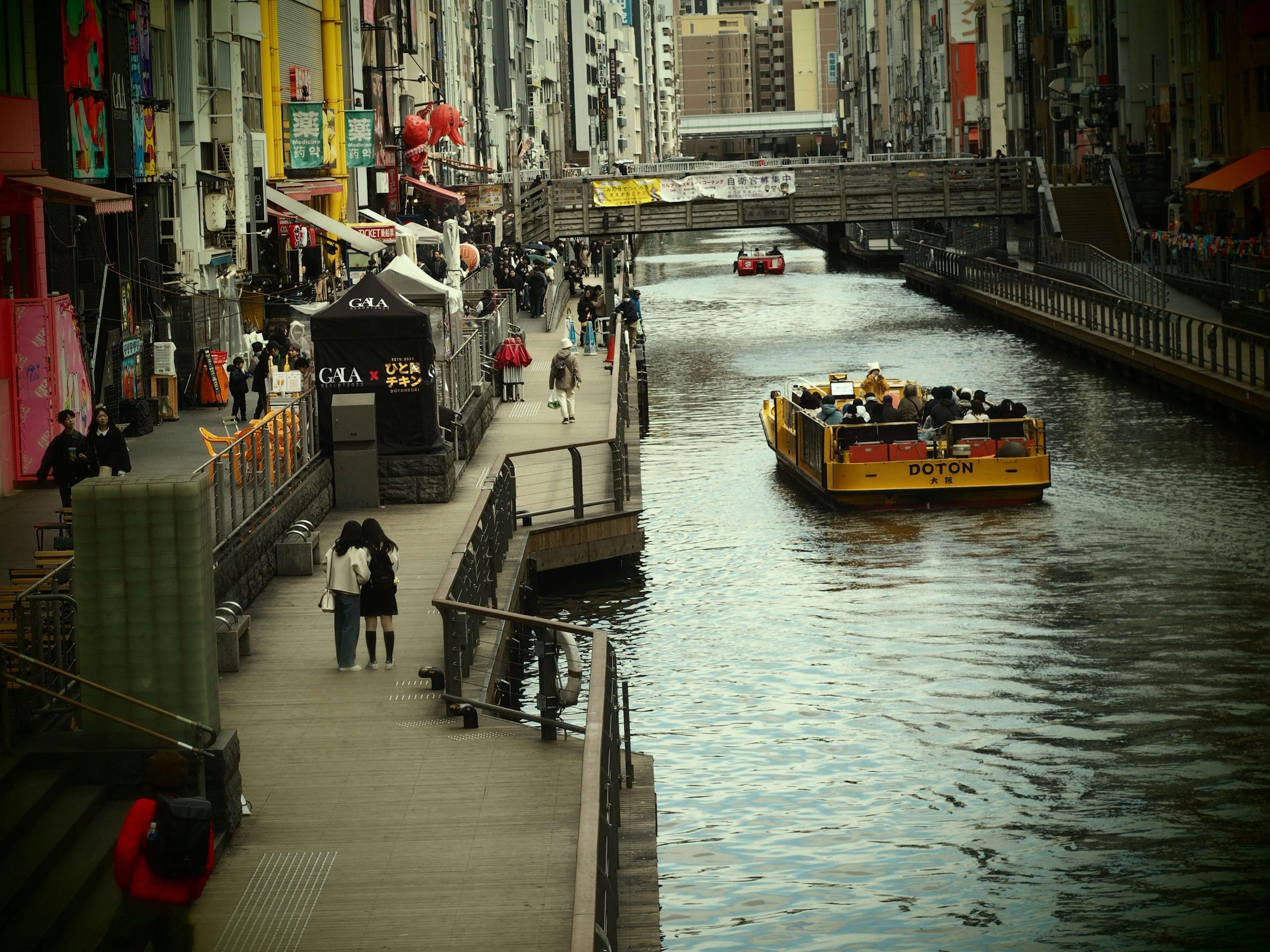 Belebter Gehweg entlang eines Kanals mit einem gelben Tourboot