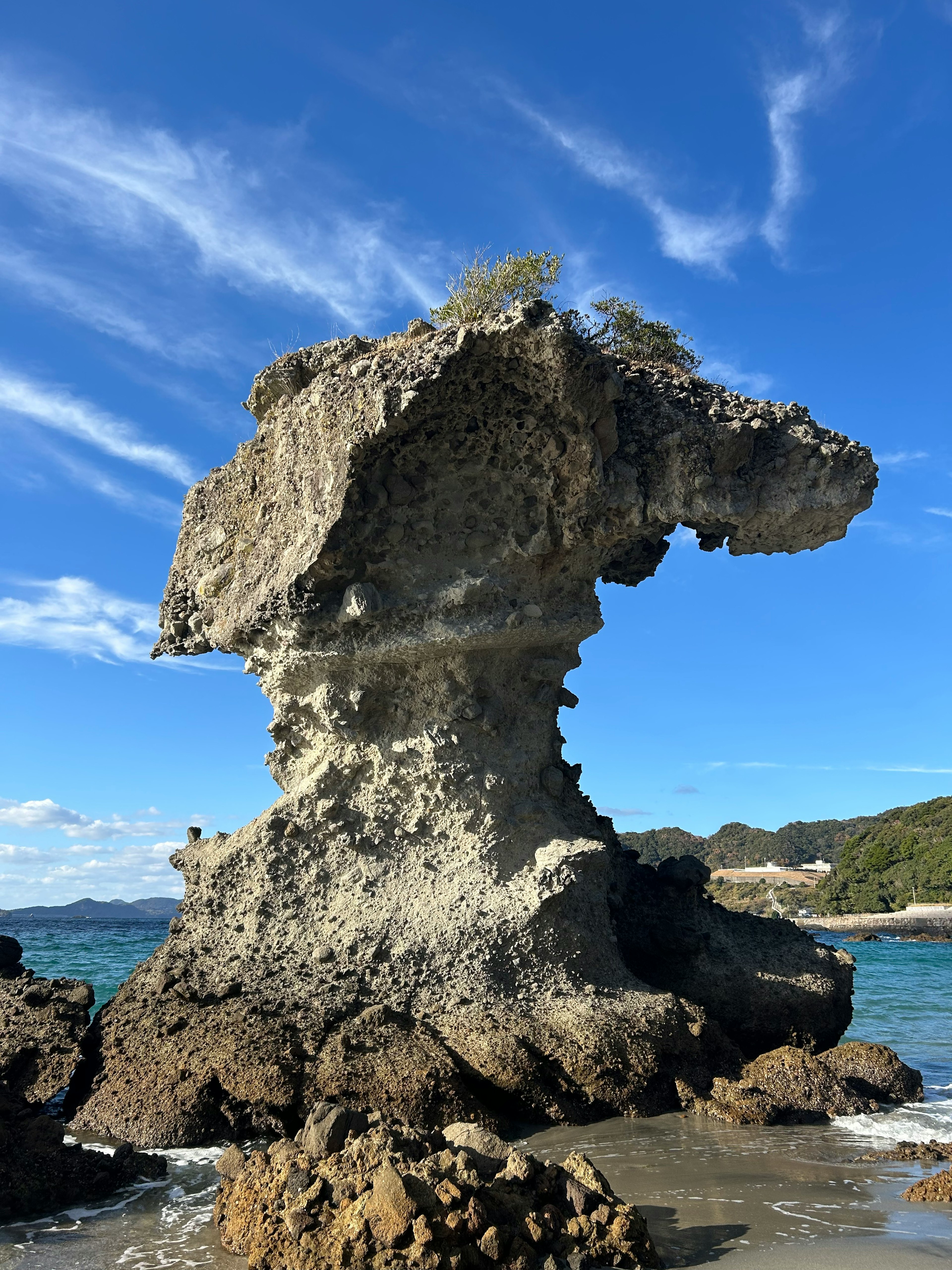 Formasi batu unik yang berdiri di pantai dengan langit biru dan laut di latar belakang