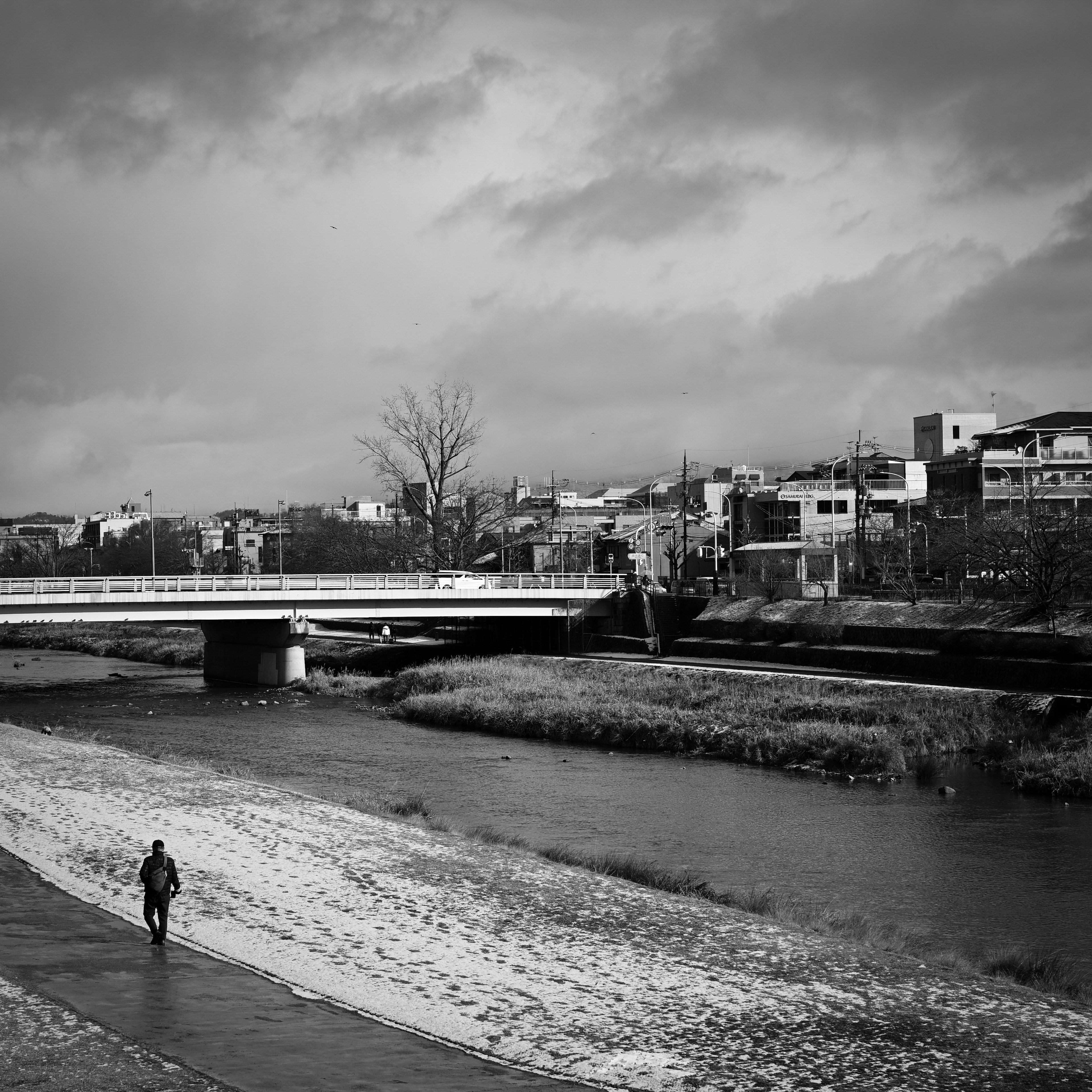 Foto en blanco y negro de un paisaje con un río y un puente una persona caminando cerca