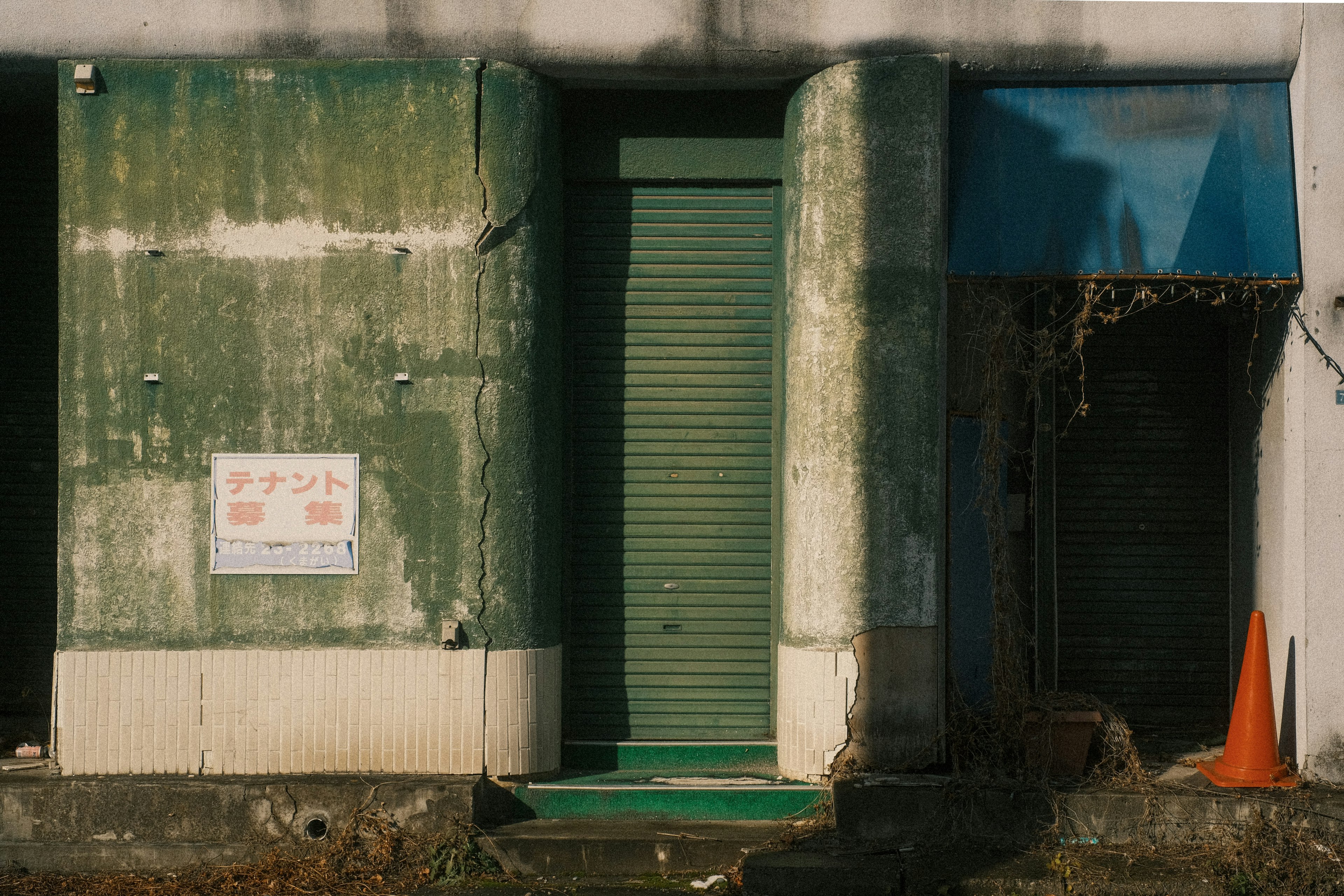 Entrada de un viejo edificio con techo azul, puerta de persiana verde, paredes desconchadas y un cono naranja