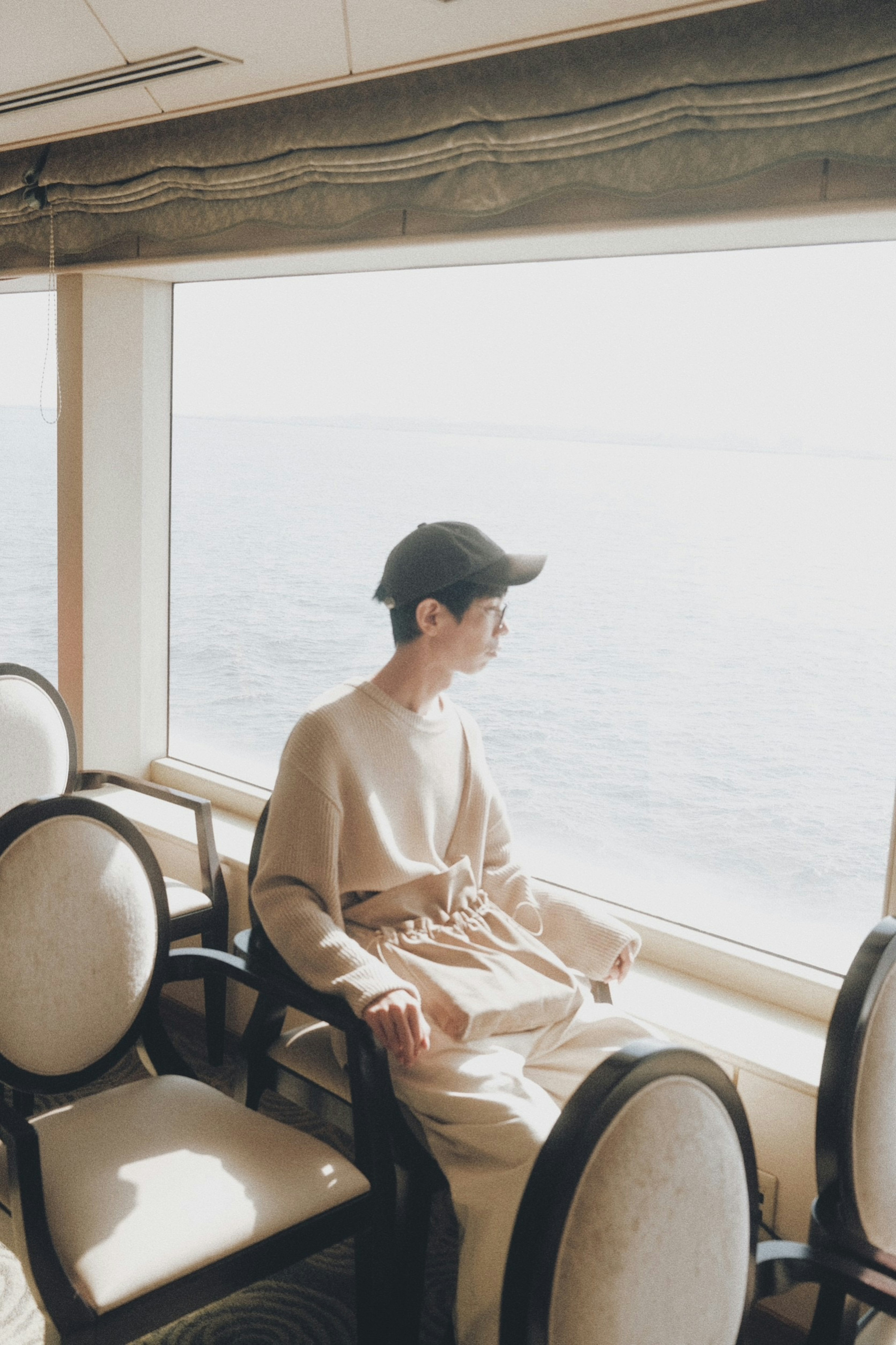 Image d'un jeune homme assis près d'une fenêtre portant un pull de couleur douce et une casquette avec une vue sur la mer calme en arrière-plan