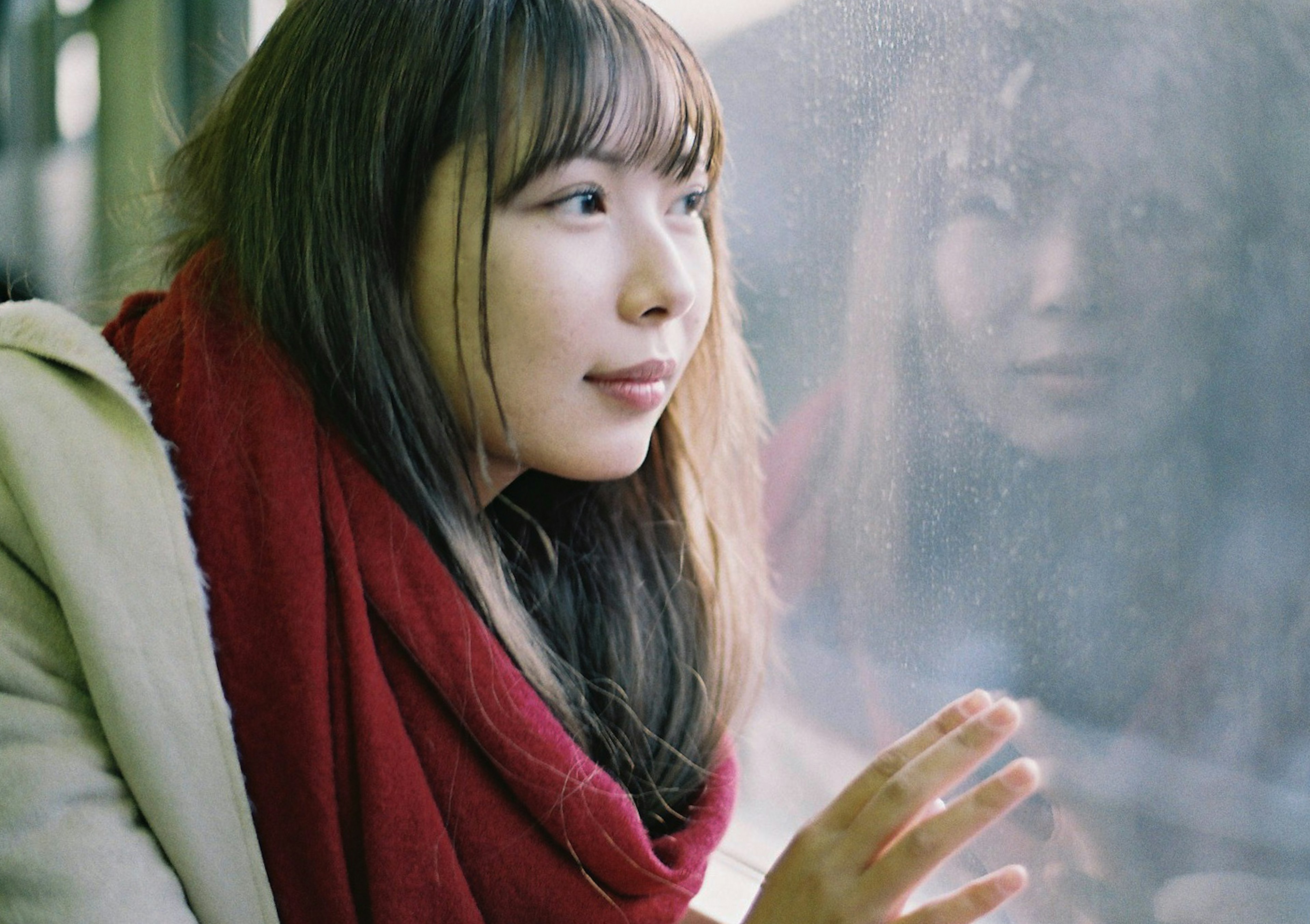 Portrait d'une femme regardant par une fenêtre, portant une écharpe rouge, avec une expression pensive et son reflet sur le verre embué