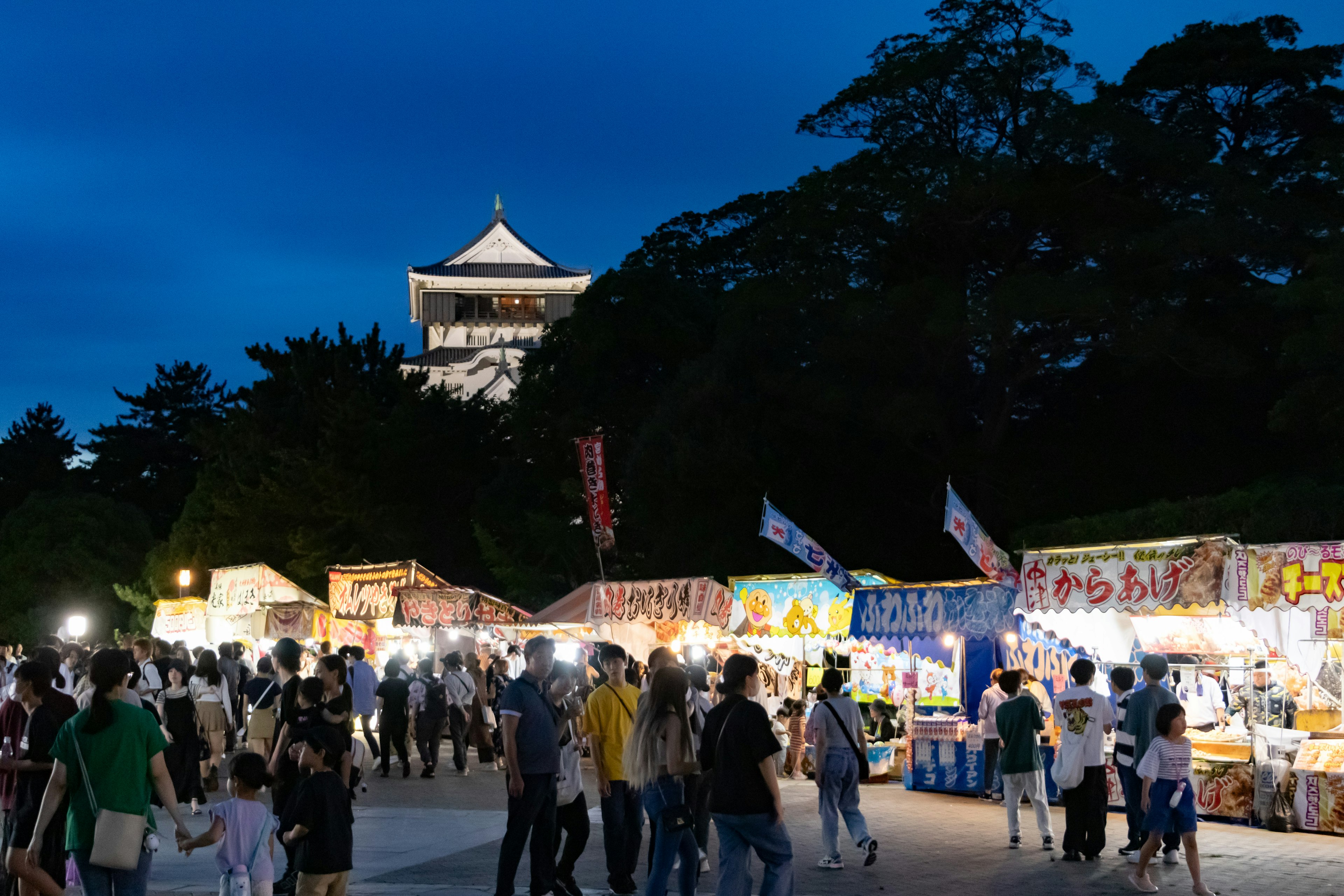 Crowd of people enjoying food stalls under a blue night sky with a castle in the background