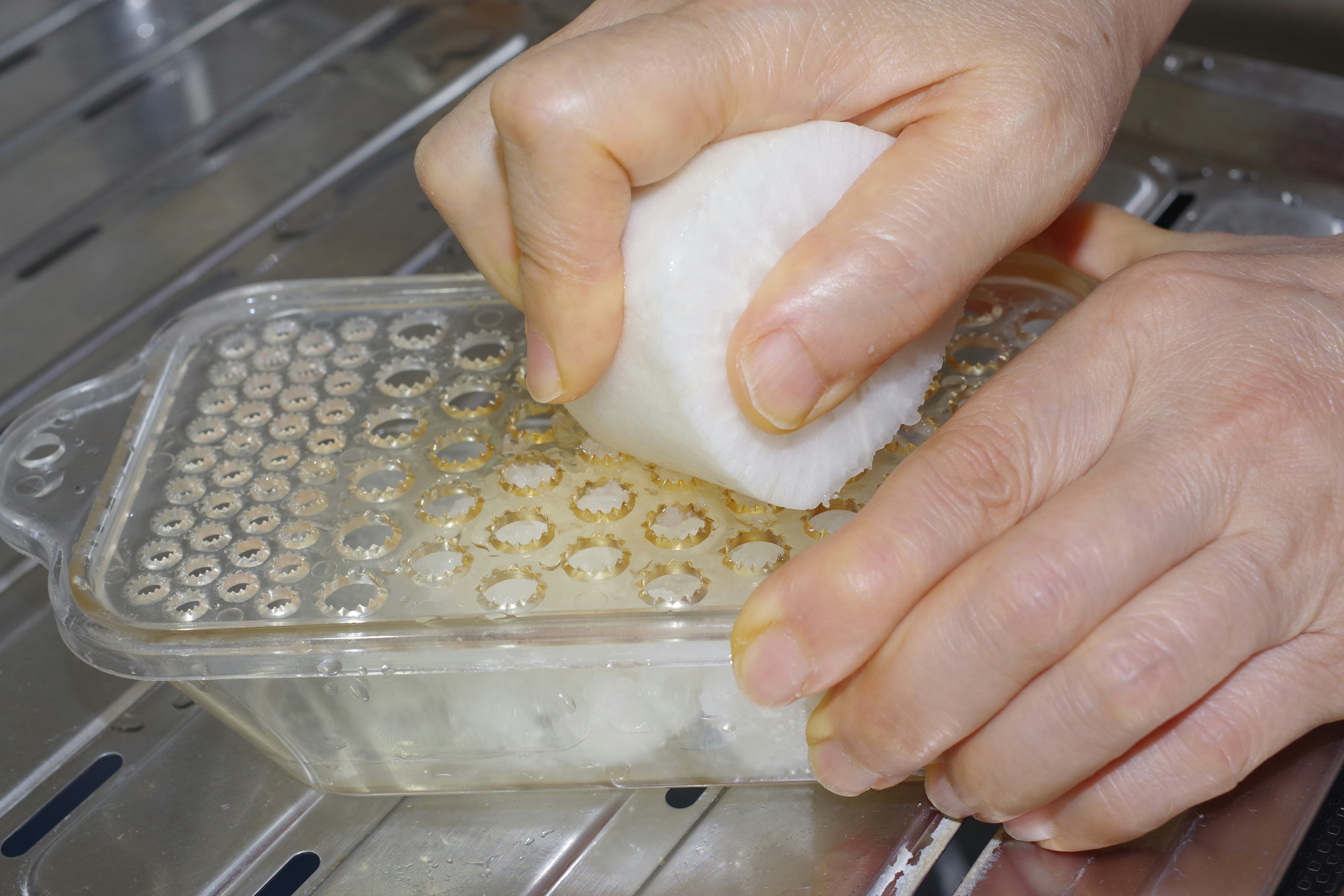 A hand grating a white object on a gray grater in a kitchen setting