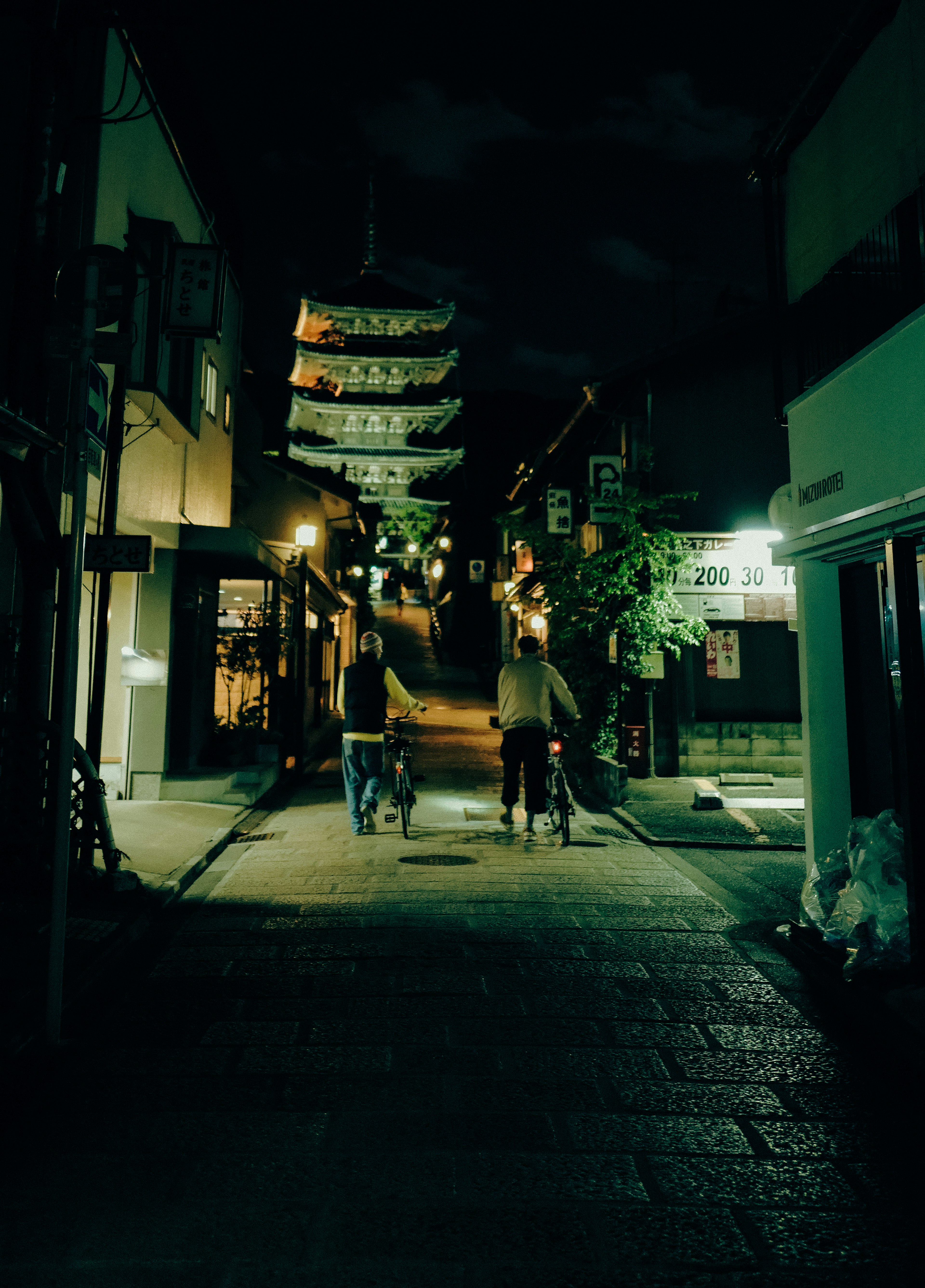 夜の街並みを自転車で進む二人の人物と提灯が見える静かな通り