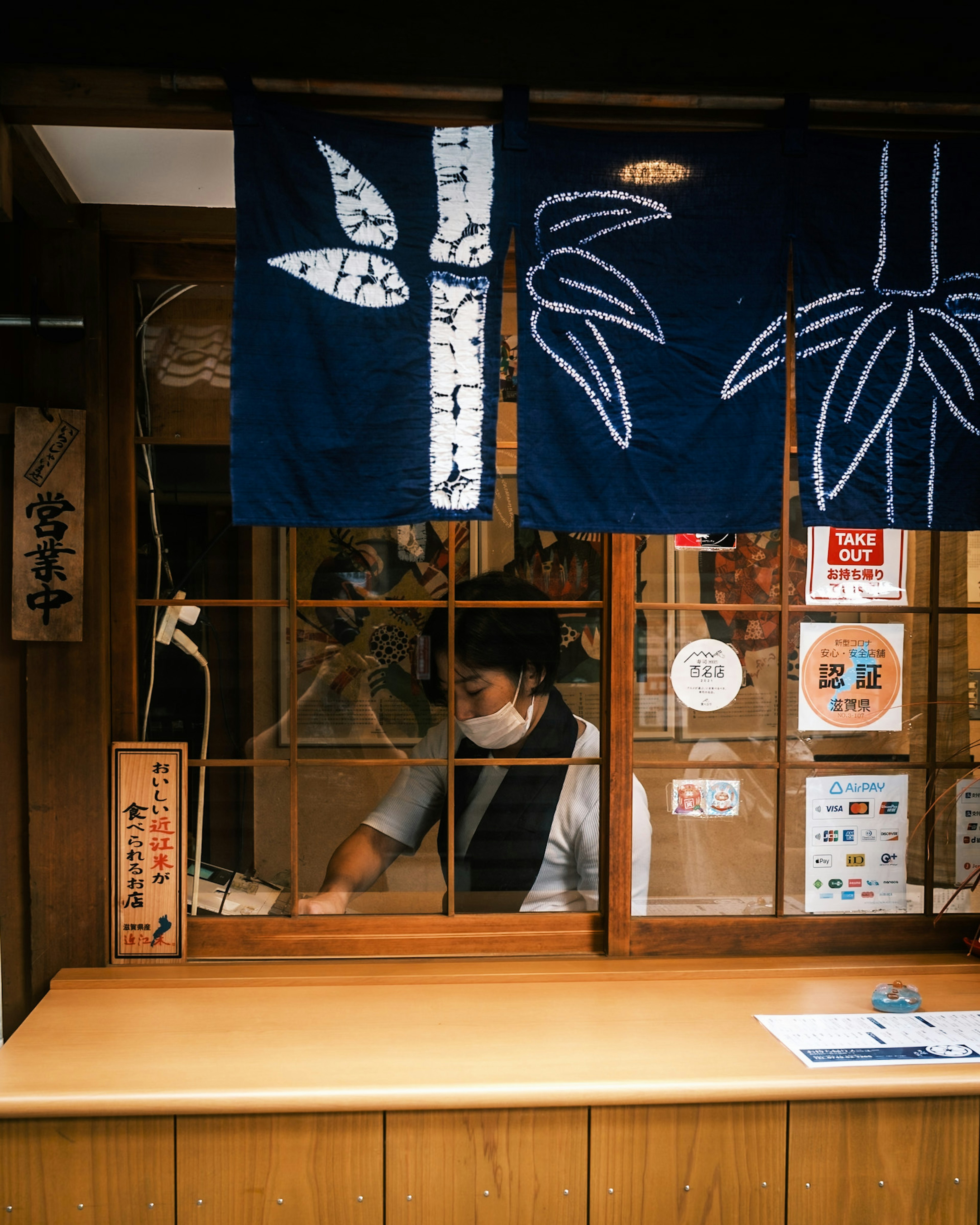 Sebuah toko Jepang tradisional dengan meja kayu dan seorang staf yang terlihat melalui jendela shoji