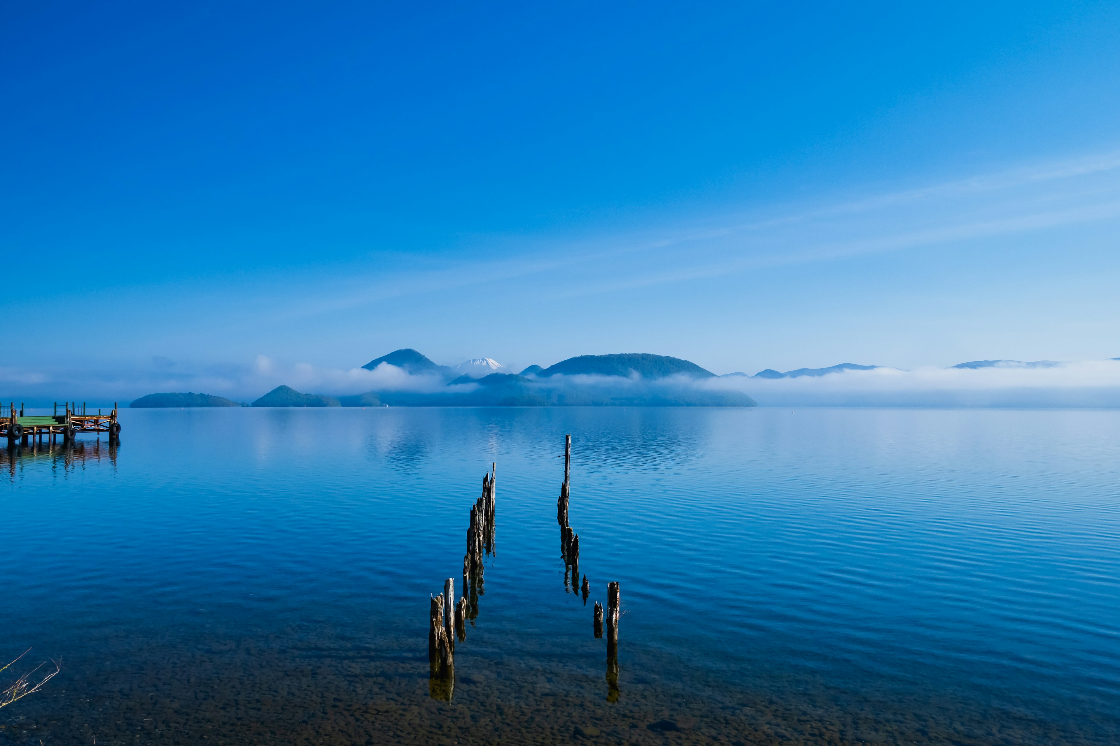 Pemandangan danau tenang dengan langit biru jernih dan pulau-pulau jauh tiang kayu menjulang dari air