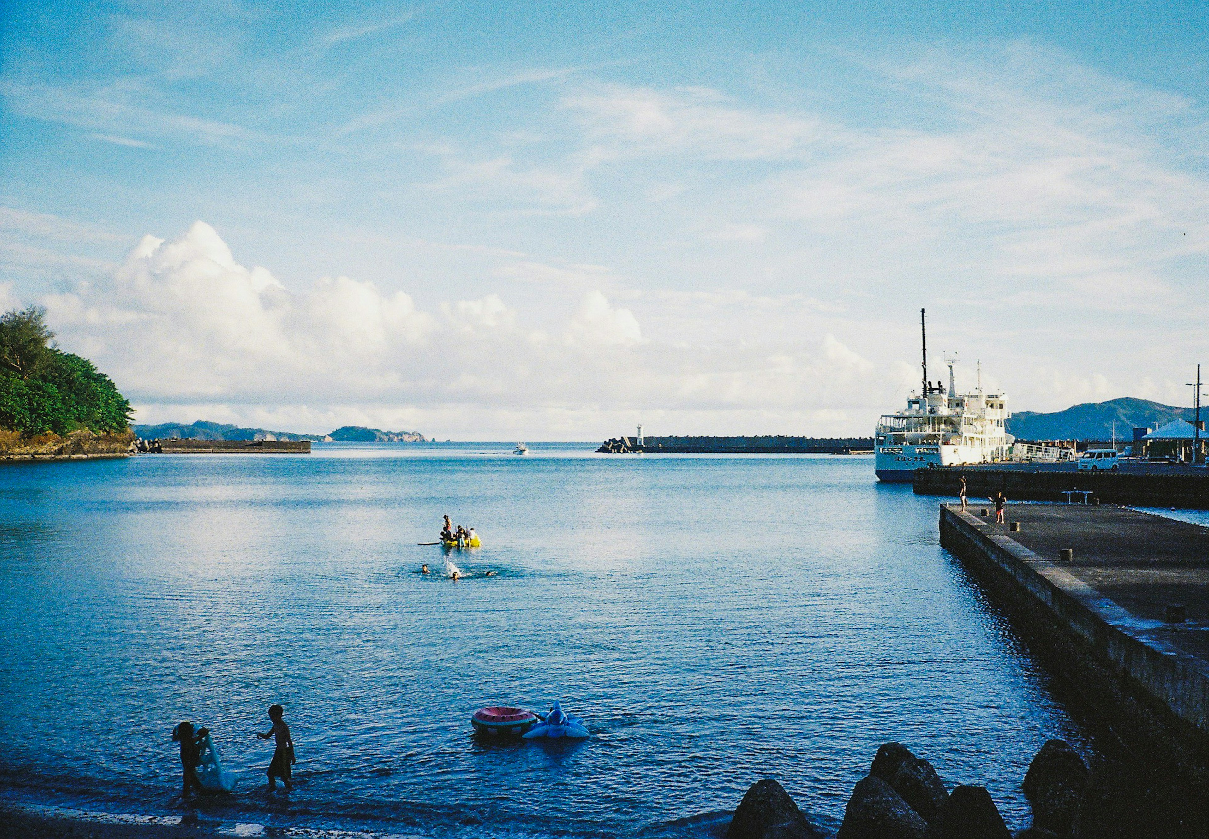 ทิวทัศน์ทะเลสงบและท้องฟ้าสีฟ้า เรือจอดอยู่ที่ท่าเรือพร้อมผู้คนเล่นที่ชายหาด