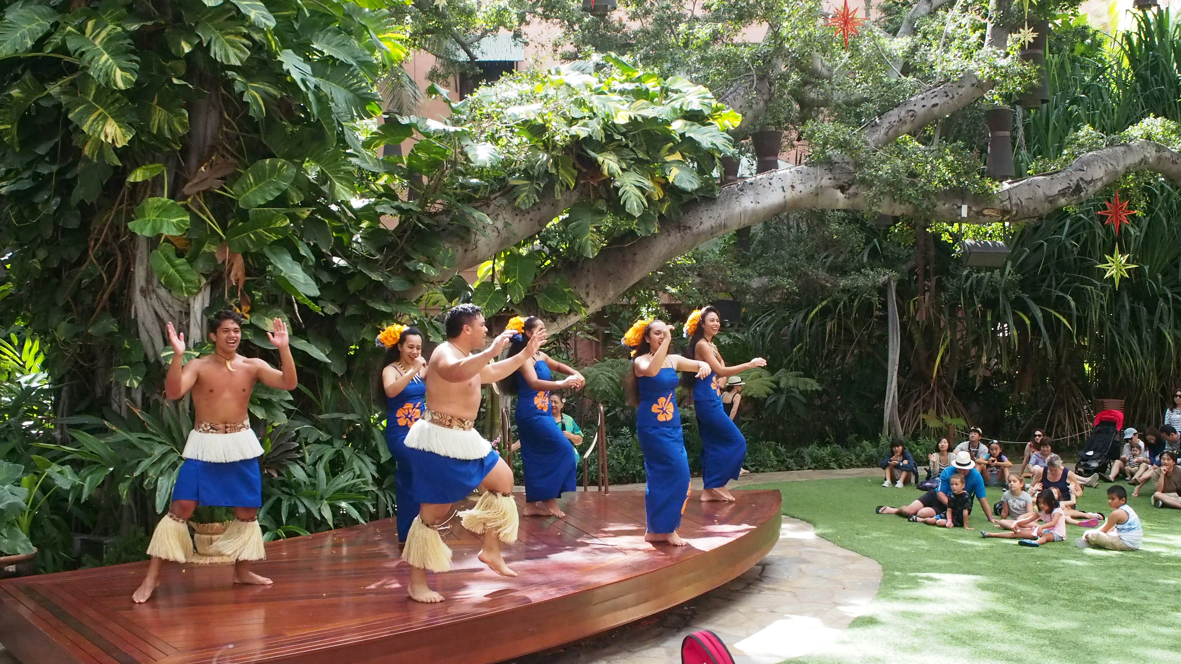 Danseurs en costumes bleus se produisant sur une scène extérieure entourée d'une végétation luxuriante et d'un public