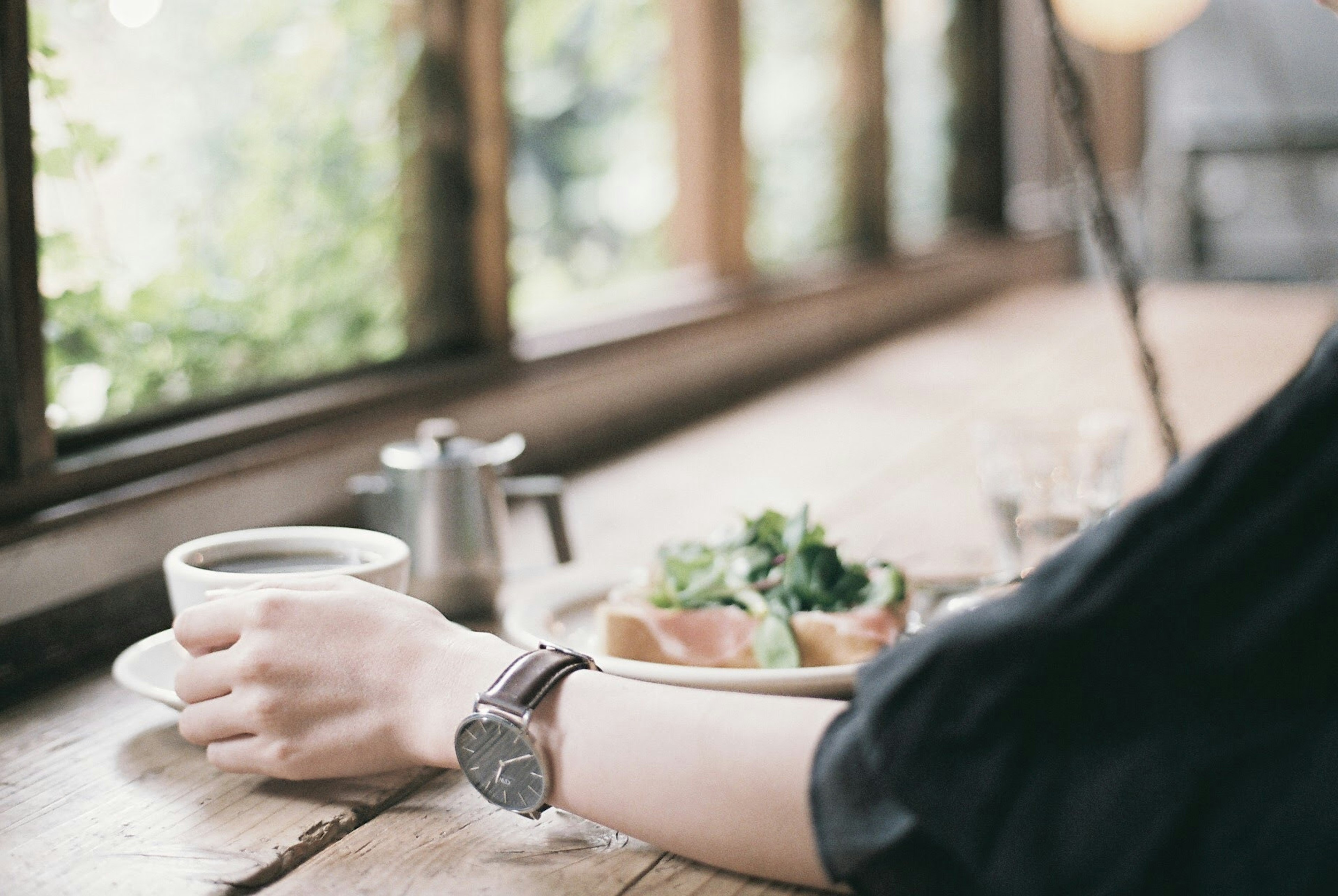 Eine Hand, die eine Tasse Kaffee nahe einem Fenster mit einem Teller Salat hält