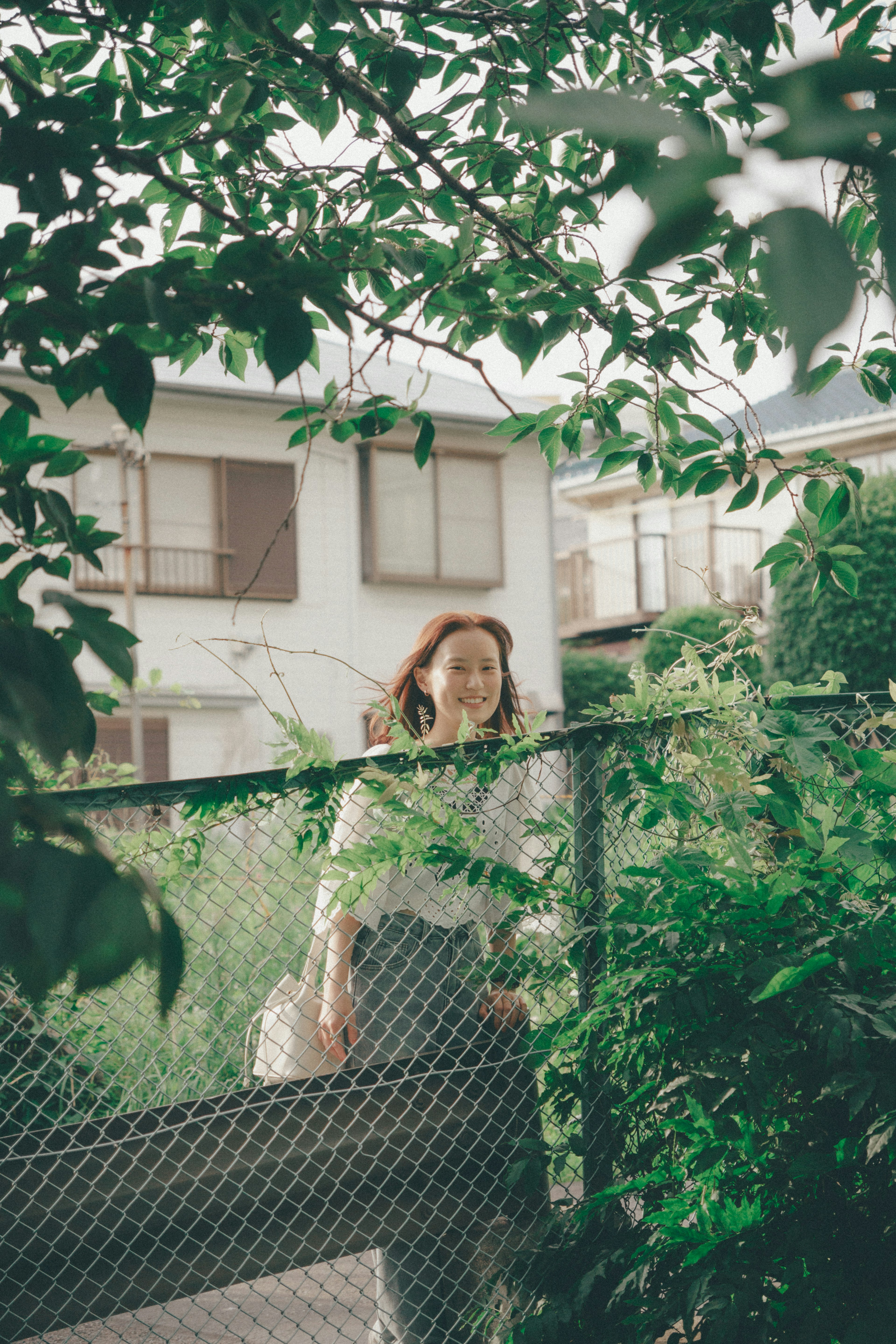 Woman standing behind a fence surrounded by green leaves and a house