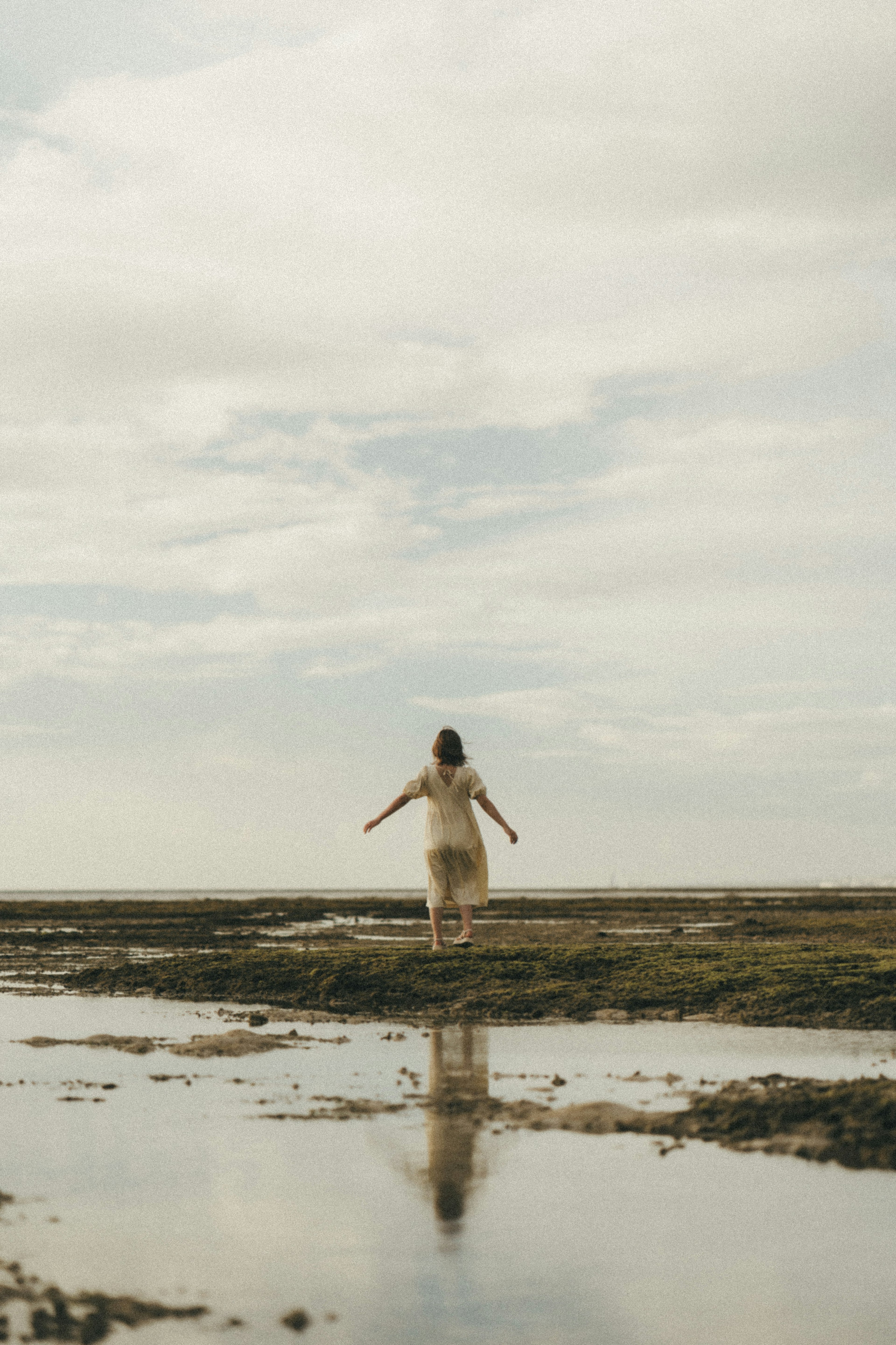 A woman standing on the beach with arms outstretched reflecting in the water and a calm sky