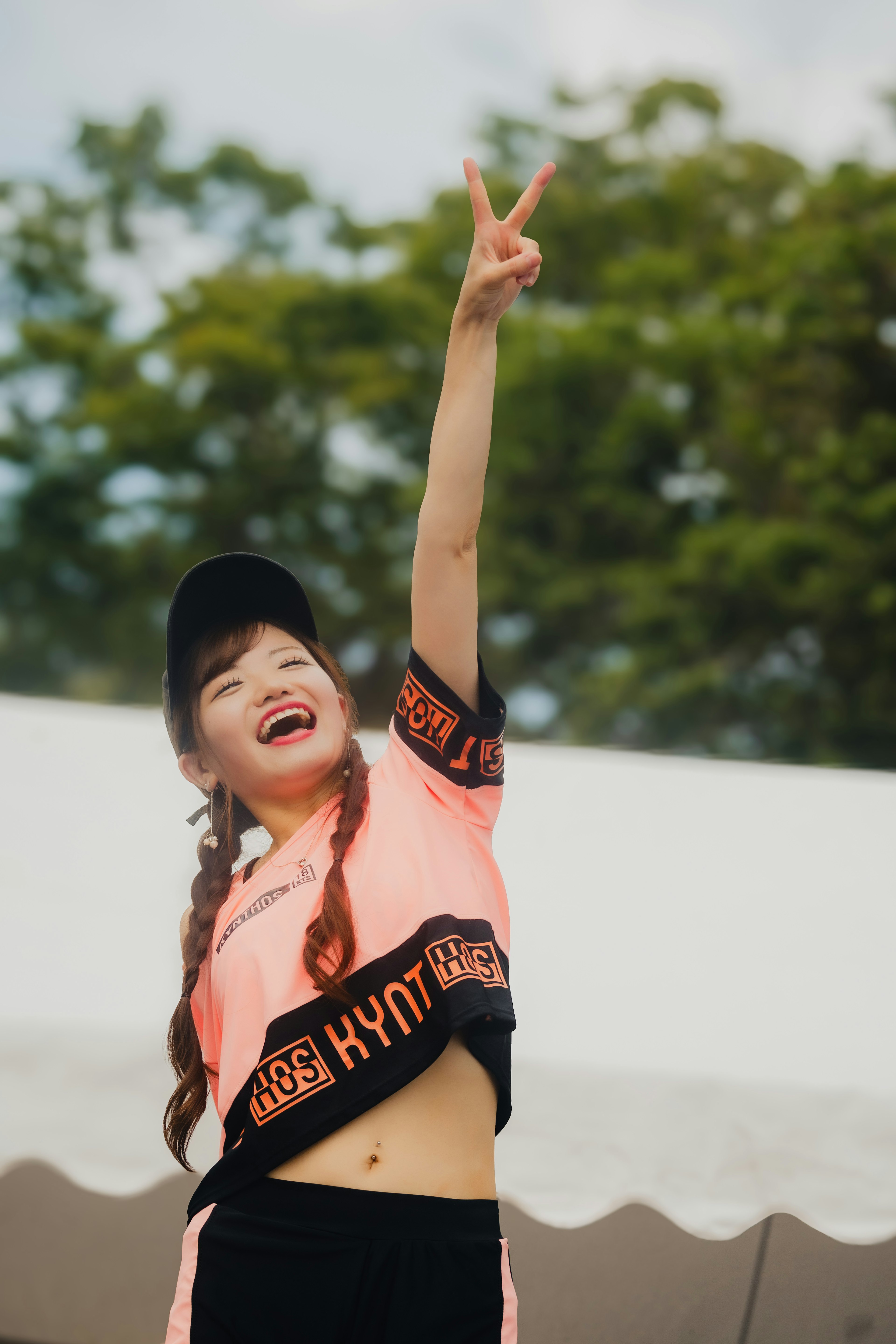 Una niña haciendo un signo de paz y sonriendo vistiendo una camiseta naranja y pantalones negros con un fondo verde