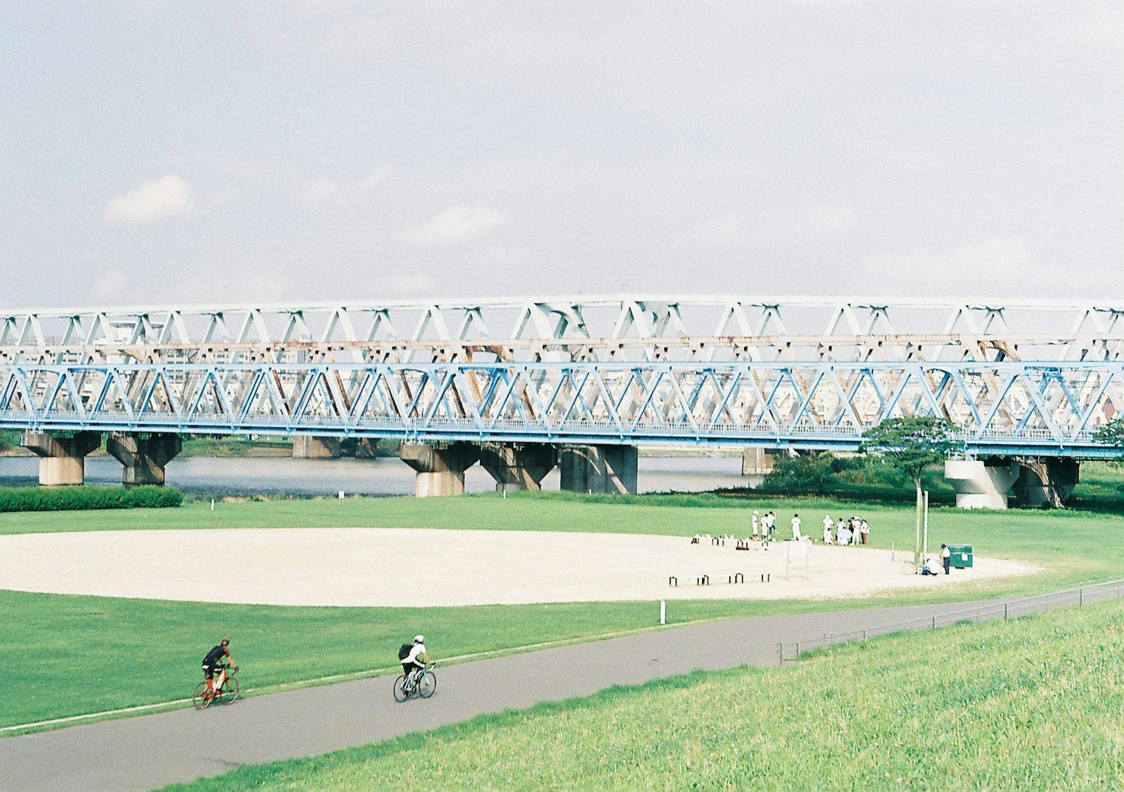 People riding bicycles under a blue bridge with a spacious green lawn