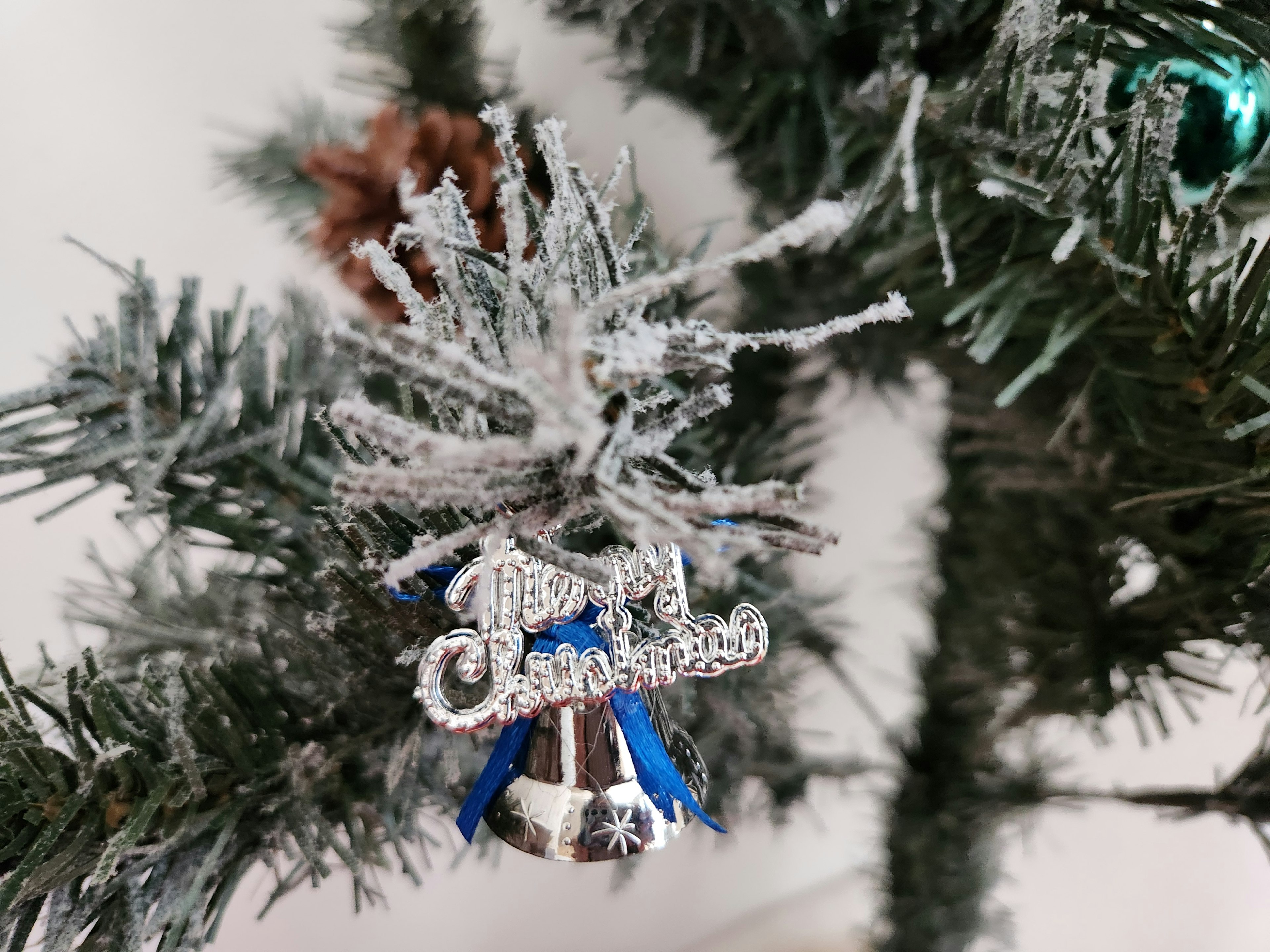 A silver bell ornament hangs from a frosted Christmas tree branch with a blue ribbon