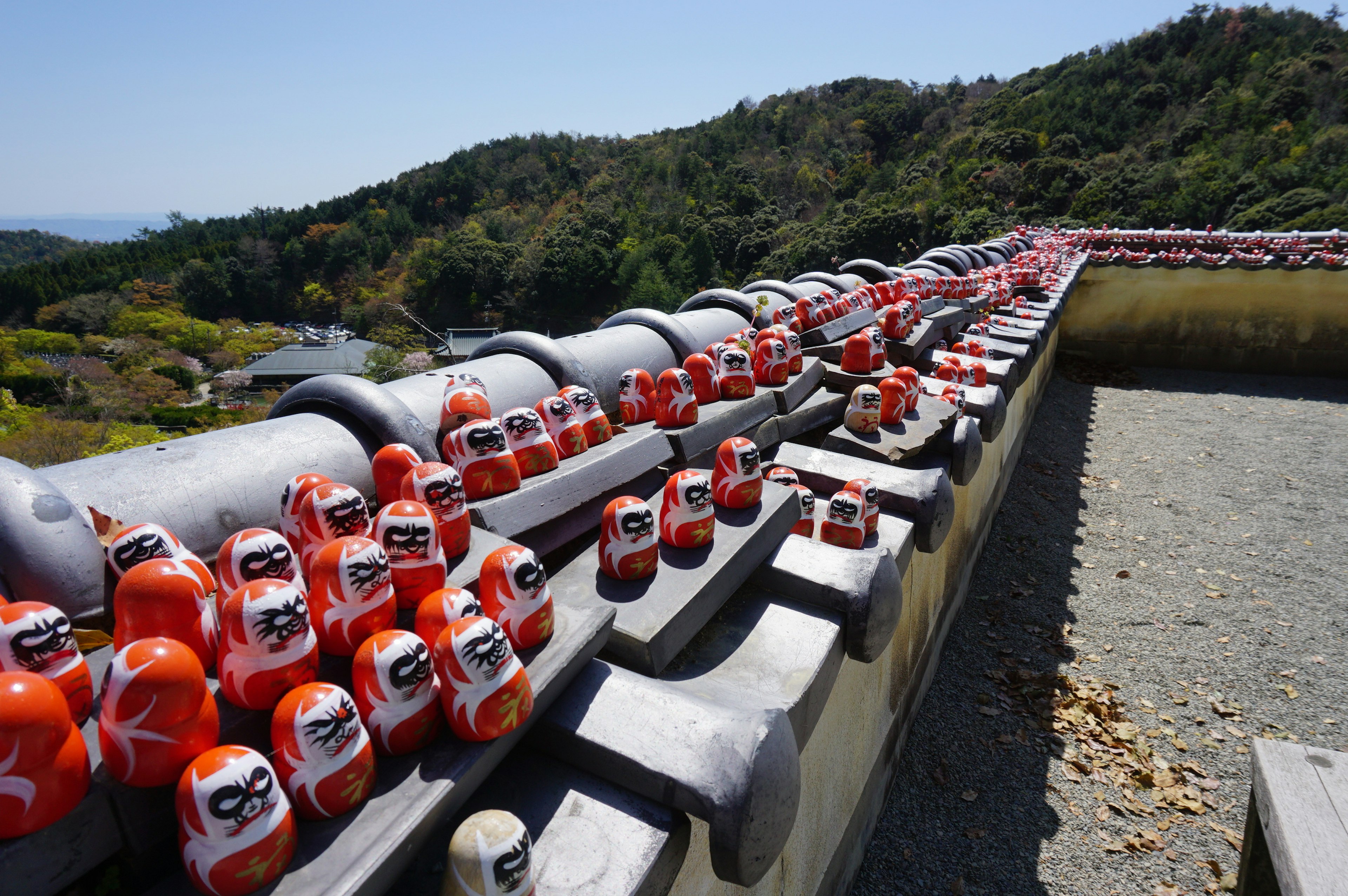 Fila de muñecas daruma rojas en un techo con montañas verdes al fondo