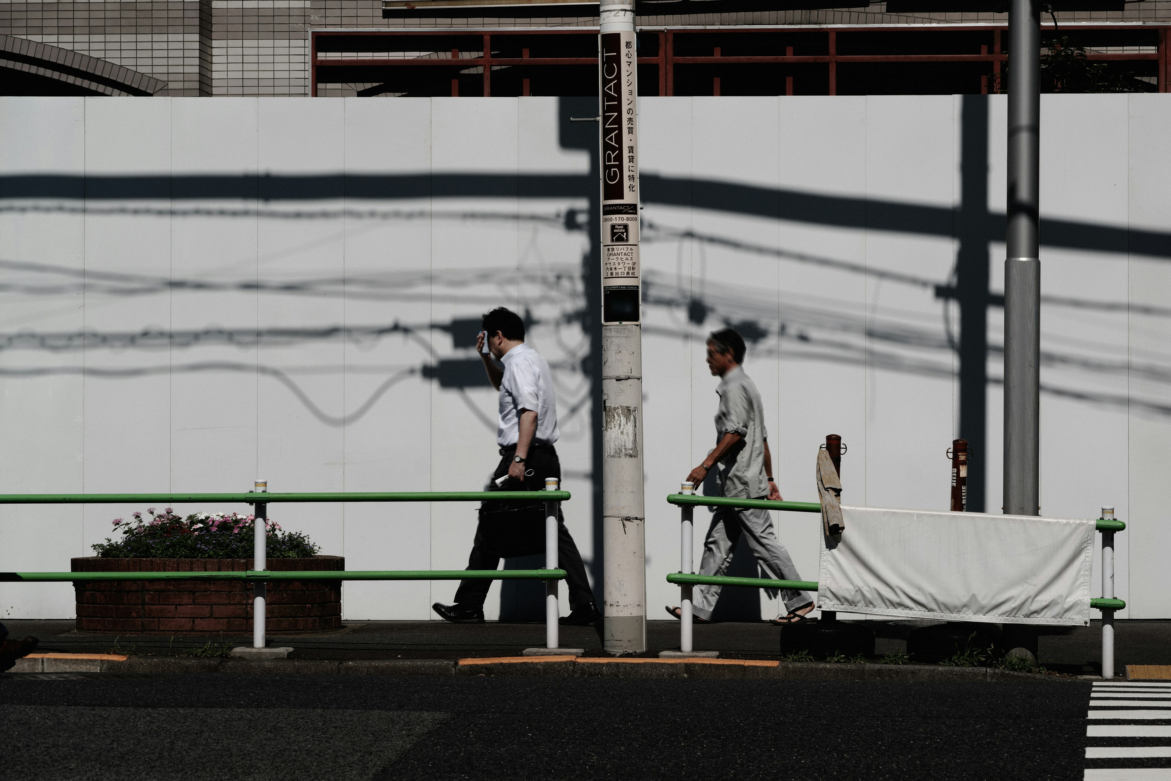 Dos hombres caminando frente a una pared blanca con sombras y plantas