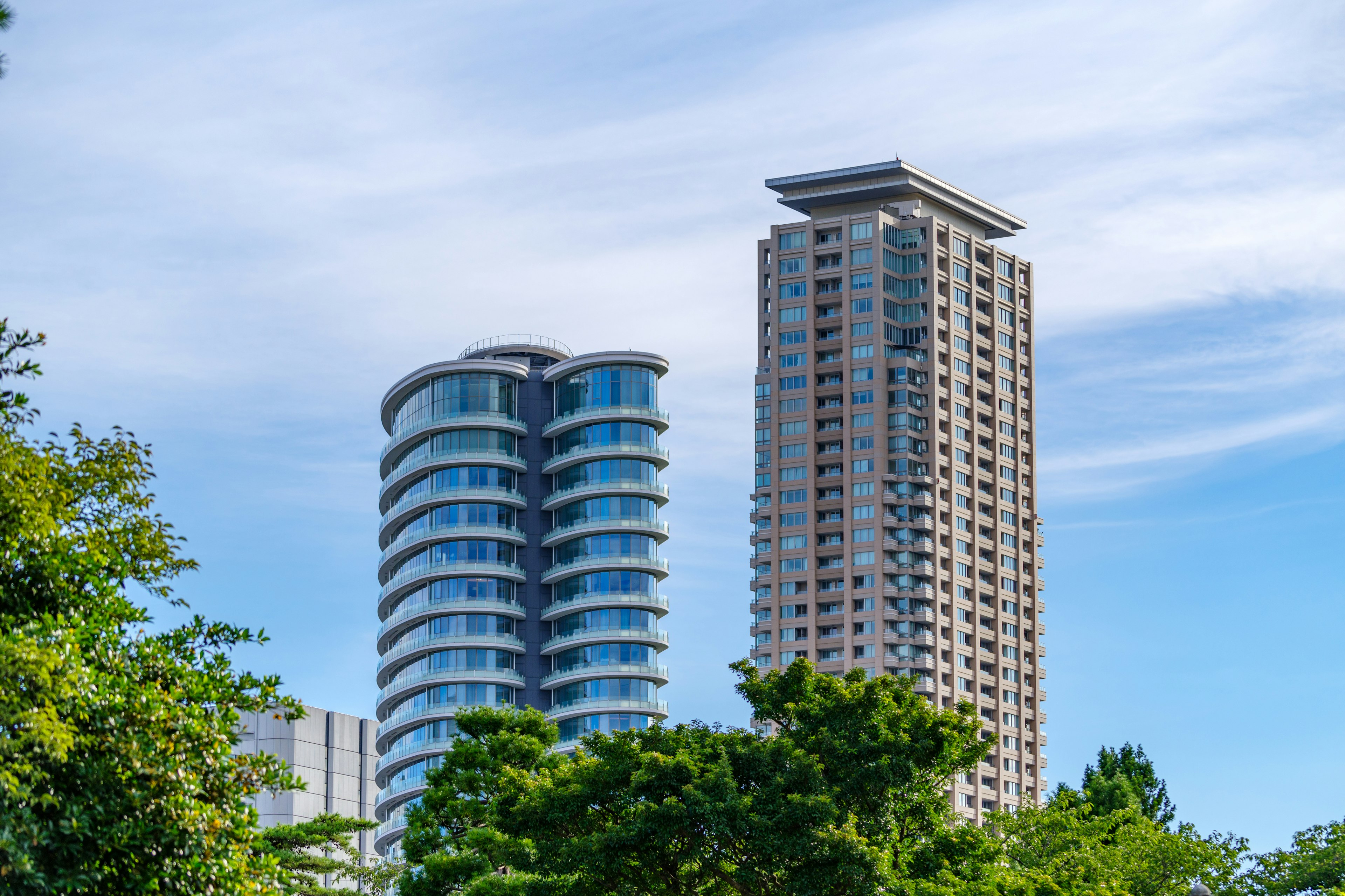 Dua gedung pencakar langit modern dikelilingi pepohonan di bawah langit biru